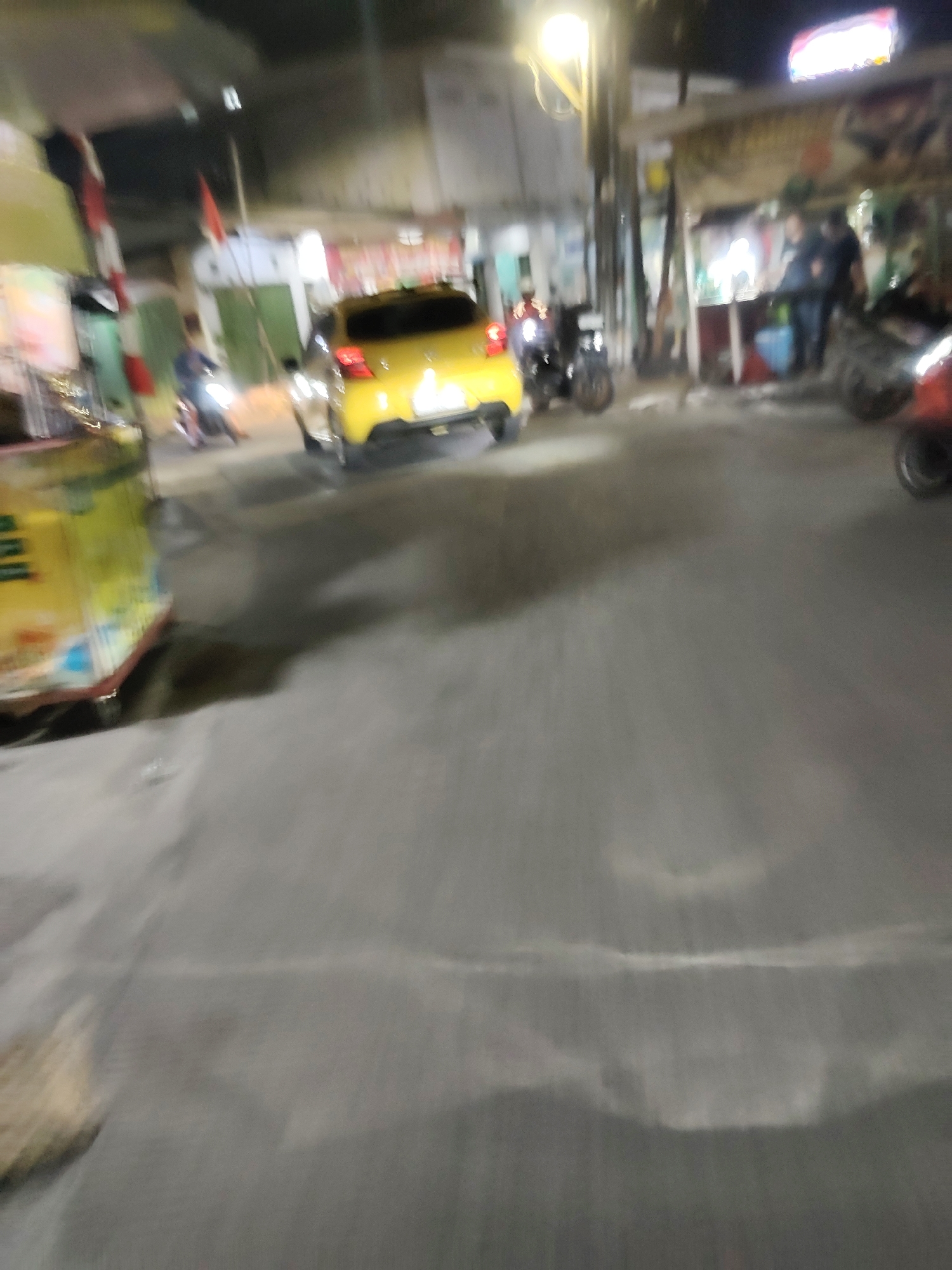 In Mekarmukti, Indonesia, at night, a yellow car is driving down a street. The street is paved and there are several shops and stalls on the side of the road. A few motorcycles are parked on the side of the street. There are a few people visible, including a person standing in one of the shops and another person on a motorcycle. The lighting in the scene is dim, with streetlights and lights from the shops illuminating the area. The image has a motion blur, suggesting that it was taken while moving.
