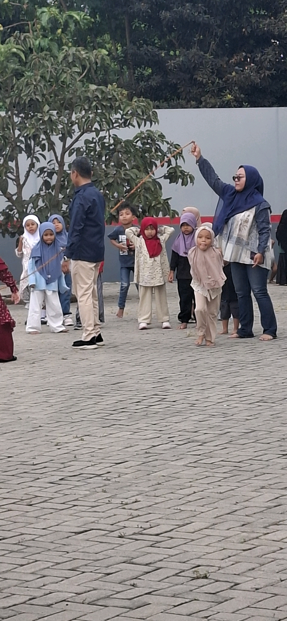 In a sunlit outdoor setting paved with grey interlocking bricks, a group of children and adults are gathered. The atmosphere suggests an informal event or outdoor activity.
The central figure is a woman wearing a dark blue hijab and glasses, dressed in a patterned blue and white top and blue jeans. She is holding a thin wooden stick upwards with her right hand, as if demonstrating or participating in an activity. Her left arm is bent at the elbow.
Surrounding her are several children. To her left, a young girl in a light brown hijab and matching loose-fitting clothing stands with her hands at her sides. Next to her is another child, also in a light brown hijab, looking forward with a slight smile. Further left, a boy in a dark blue t-shirt and blue jeans stands with his hands on his hips.
To the right of the woman with the stick, a child in a red hijab and a patterned cream-colored outfit stands with hands behind their head. Behind this child, another child in a purple hijab is partially visible.
To the left of the frame, a man in a navy blue collared shirt and light beige pants stands facing away from the camera, appearing to observe the scene. Several other children are visible in the background, some wearing hijabs in white and light blue. The ground is a patterned brick pavement, and in the background, there are trees with lush green foliage and a light-colored wall. The overall impression is of a community gathering or an educational activity taking place in a public area. The presence of both adults and children, along with the casual attire, suggests a relaxed and engaging environment. The location is identified as the city of Blitar, Indonesia, implying a cultural context where head coverings like hijabs are common.