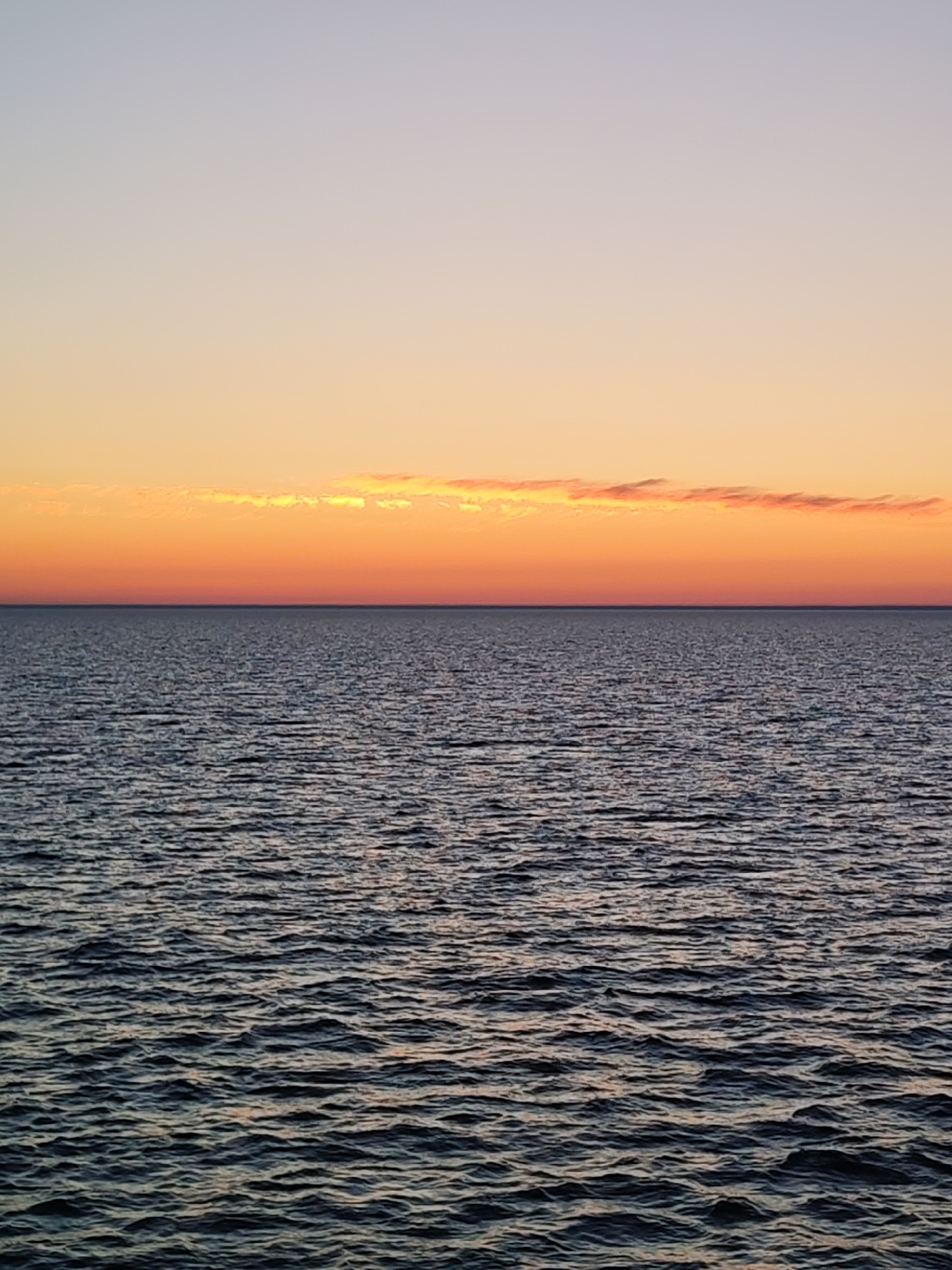 The image shows a body of water with small waves, likely an ocean or large lake, under a sky transitioning from light orange near the horizon to a light purple-grey further up. A thin strip of orange clouds sits just above the horizon. The location and city are unknown, and there are no people or specific objects visible in the frame beyond the water and sky.