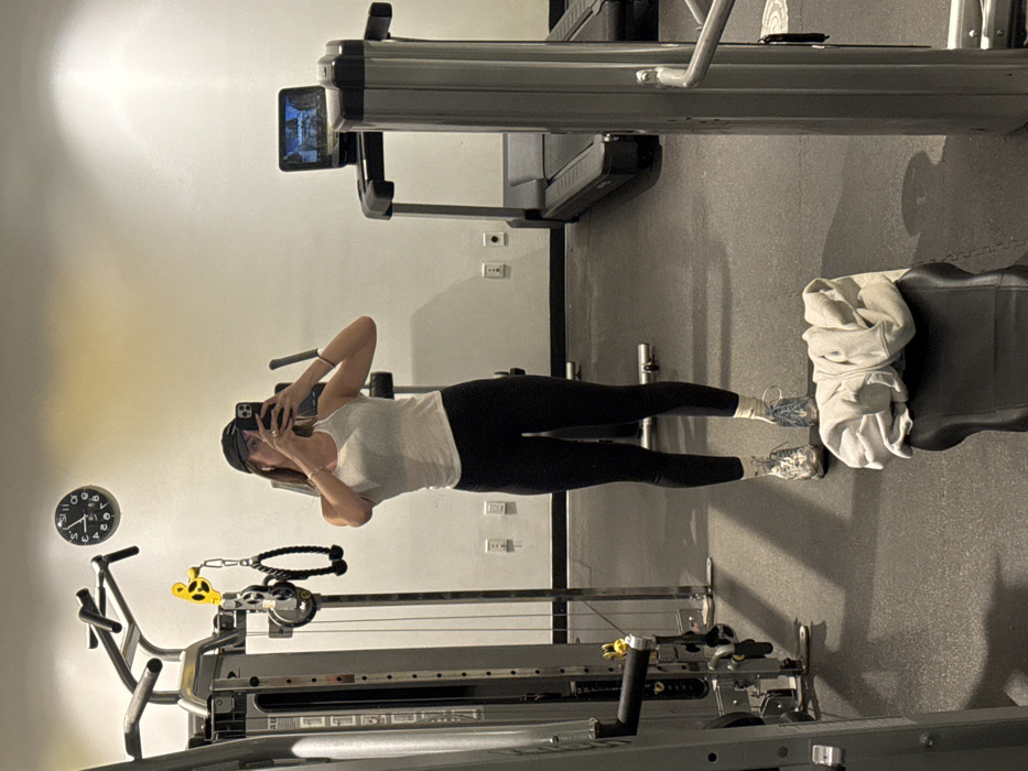 A woman in a white tank top, black leggings, and silver sneakers takes a photo of herself in a gym. She stands between a weight machine and a treadmill. A folded, light-colored hoodie rests on a black bench to her right. A black clock hangs on the wall behind her. Electrical outlets are visible along the lower portion of the wall.