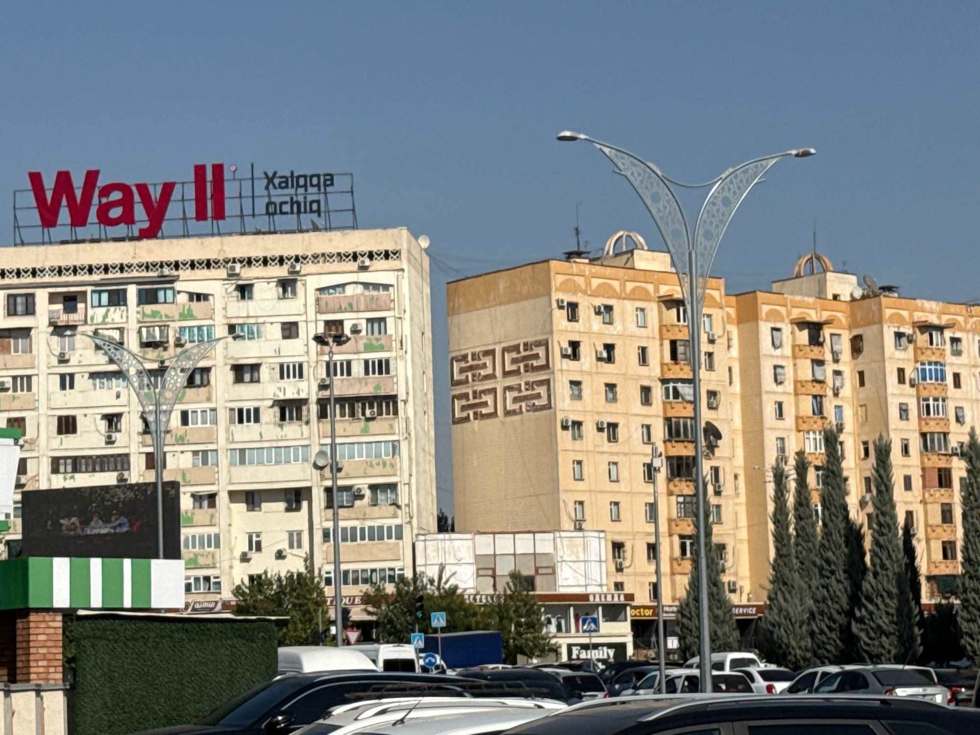 This is a daytime, eye-level shot of a city street in Tashkent, Uzbekistan. In the foreground, the tops of several cars are visible. Behind the cars, a bright green hedge runs horizontally across the image. Above the hedge, a large, dark LED screen displays an image of people seated around a table. To the left of the screen is a white and green striped structure. Behind this, a tall, light-colored apartment building with numerous windows and air conditioning units is visible. On top of this building, a large red sign reads "Way II" with smaller text to the right. Further back, a light yellow apartment building with similar architectural features stands. A distinctive, ornate street lamp with a decorative metal design is positioned in the middle ground. More apartment buildings, cypress trees, and street signs are visible in the background. The sky is clear and blue.