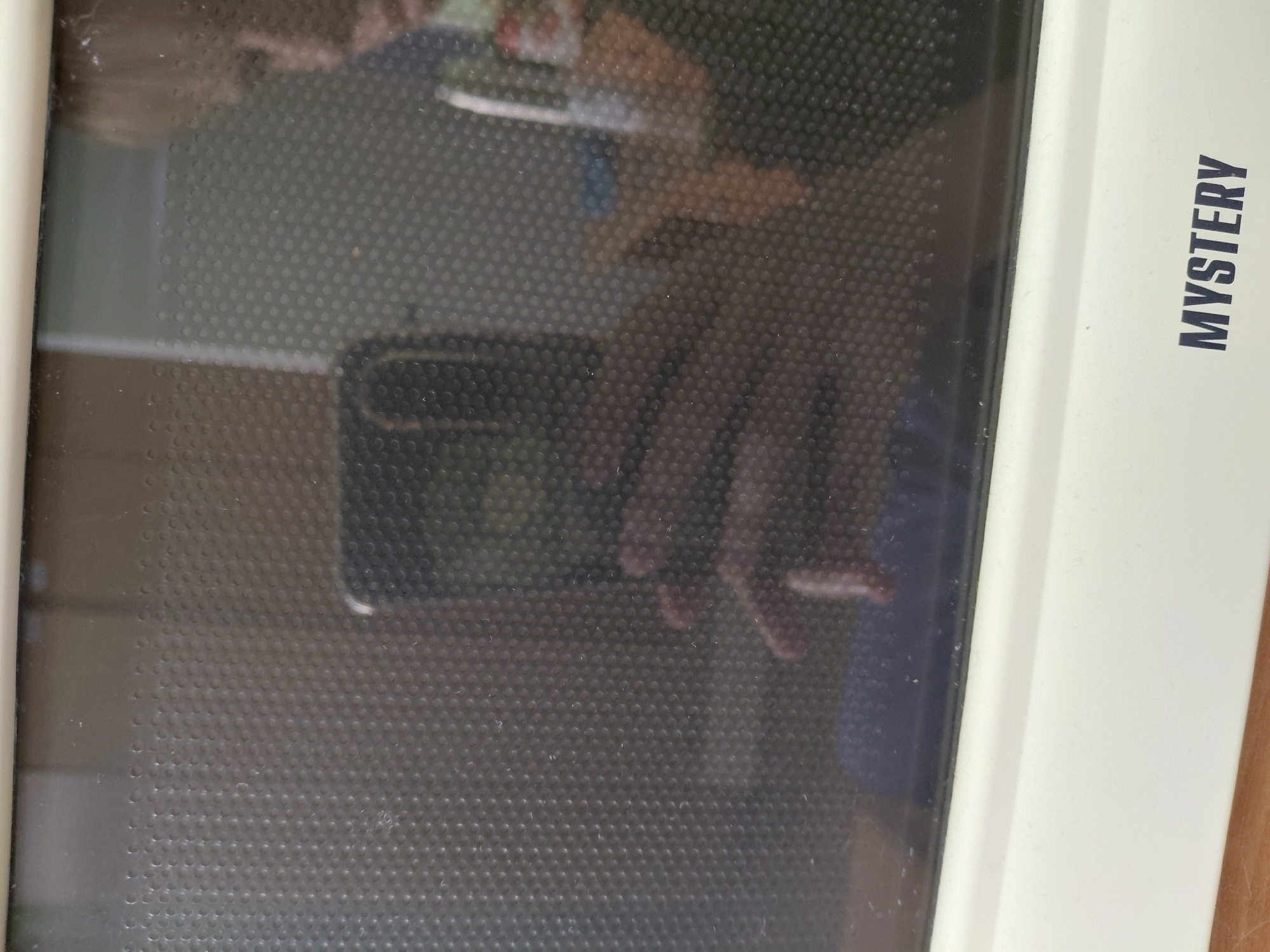 A close-up, slightly angled shot captures the front of a light-colored microwave oven. The microwave's door is dark and highly reflective, featuring a fine, uniform mesh or dot pattern. Prominent reflections are visible on the door, showing what appears to be a person's hands and fingers holding a dark rectangular object (possibly a smartphone) as if taking the picture. Other less distinct reflections of the person's upper body and head are also visible, along with parts of the surrounding indoor environment, likely a kitchen with walls or cabinets.
On the right side of the microwave, oriented vertically, the brand name "MYSTERY" is clearly visible in dark, capitalized letters on the light-colored frame. The microwave itself appears to be white or off-white. To the right of the microwave, a wooden surface, possibly a countertop or table, is visible. The surfaces of the microwave door and frame show some dust or small particles.
The image depicts a moment of someone photographing the microwave, with the camera's angle and reflections being key elements of the composition. The location context provided is Nadym, Russia, although this is not visually evident in the image.