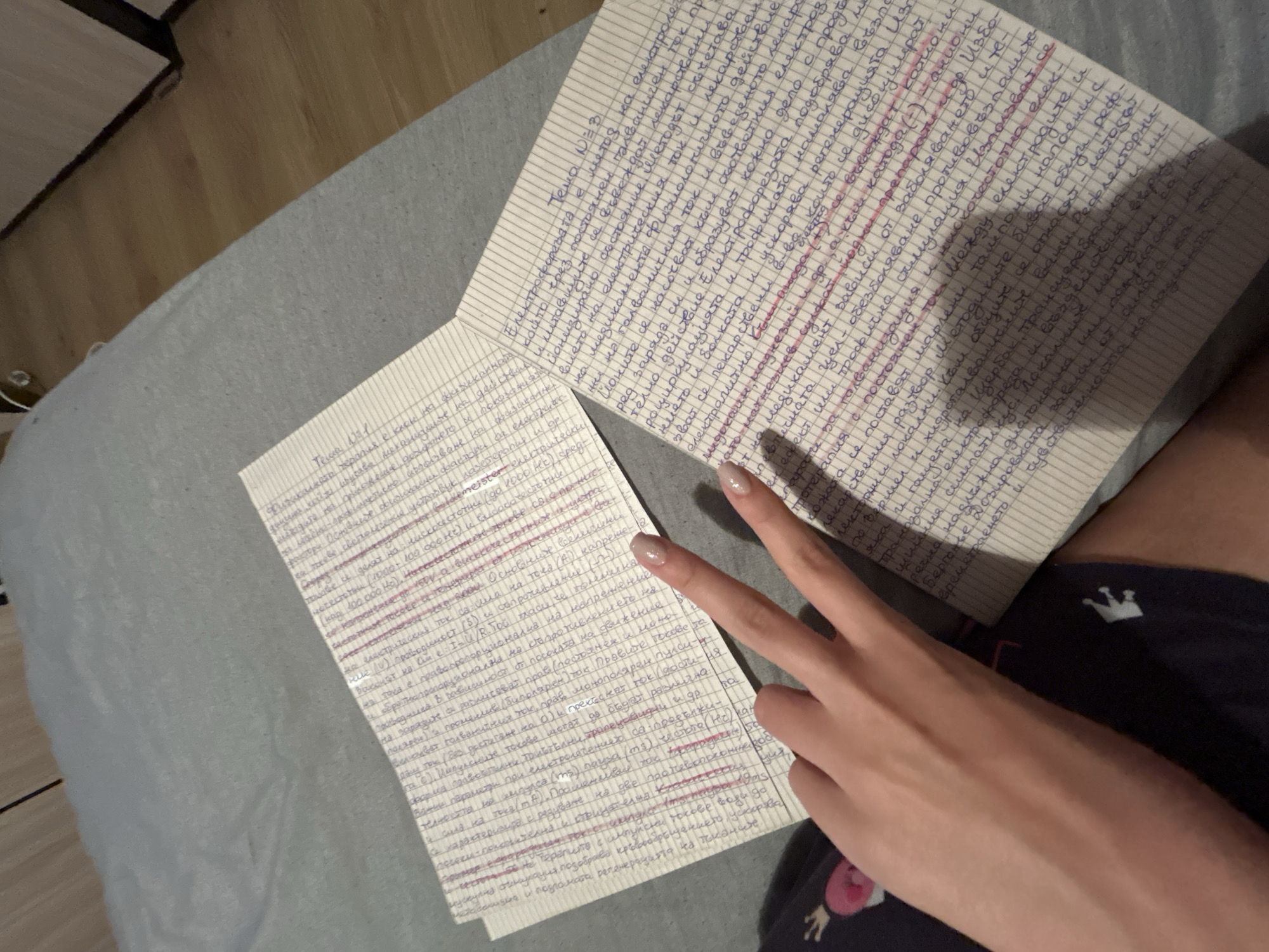 A person is sitting on a bed, holding two sheets of paper with handwritten notes. The person's left hand is raised, forming a peace sign. The sheets of paper are filled with dense, illegible handwriting in blue ink on lined paper. The person is wearing dark-colored shorts with a white crown pattern. The bed has a light gray cover. The background includes a wooden floor and a partially visible shelf or cabinet.
