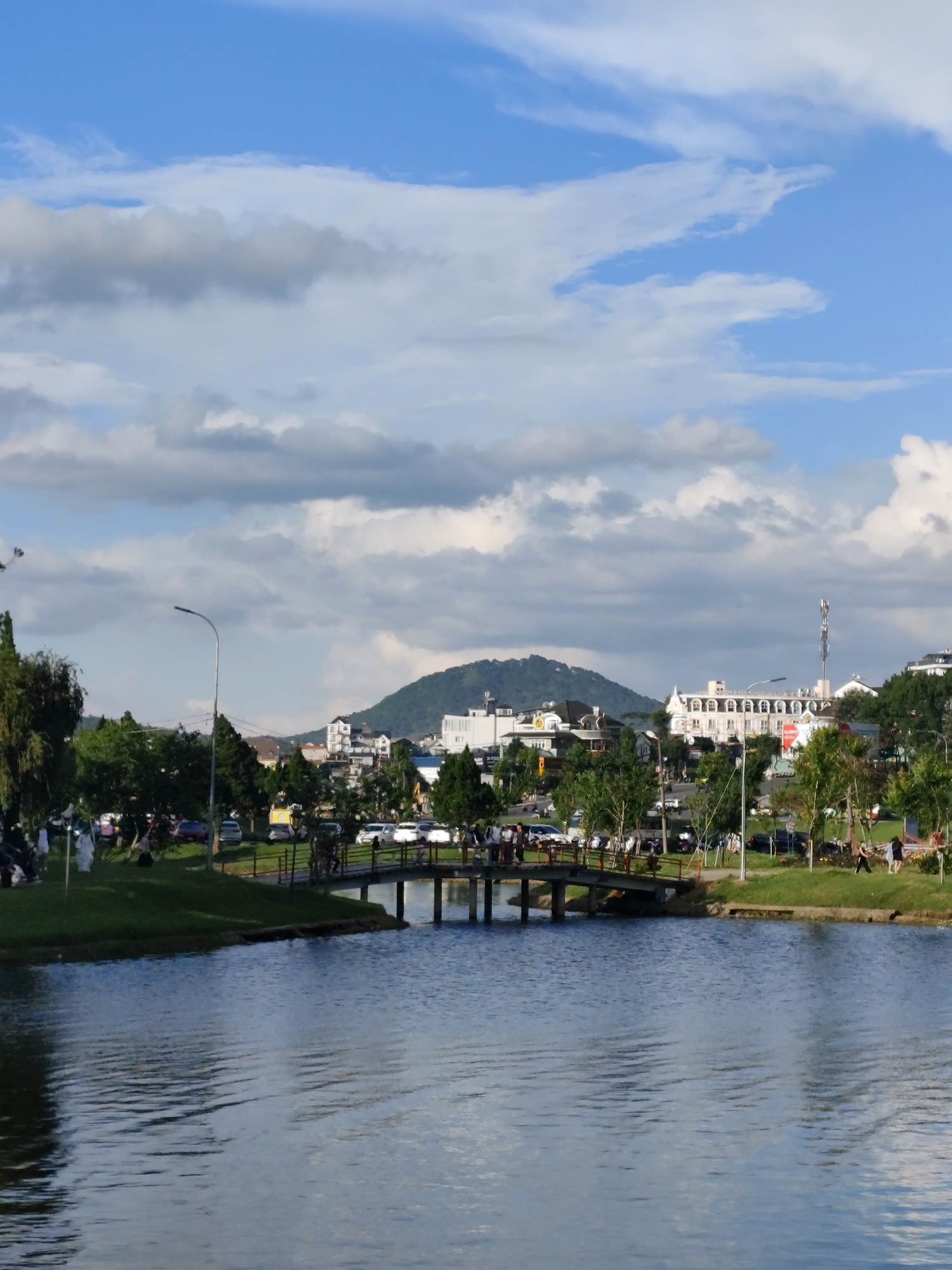 The image depicts Xuan Huong Lake in Da Lat, Vietnam, on a partly cloudy day. The foreground is dominated by the calm, blue water of the lake. A concrete bridge with railings spans across a section of the lake, with people visible walking on it. The middle ground features a grassy bank with trees and a street lined with parked cars and buildings. In the background, a lush green hill with a town built on its slope rises, topped by a few buildings and a telecommunications tower. The sky above is a vibrant blue with scattered white and gray clouds. The overall scene is one of a peaceful urban park setting.