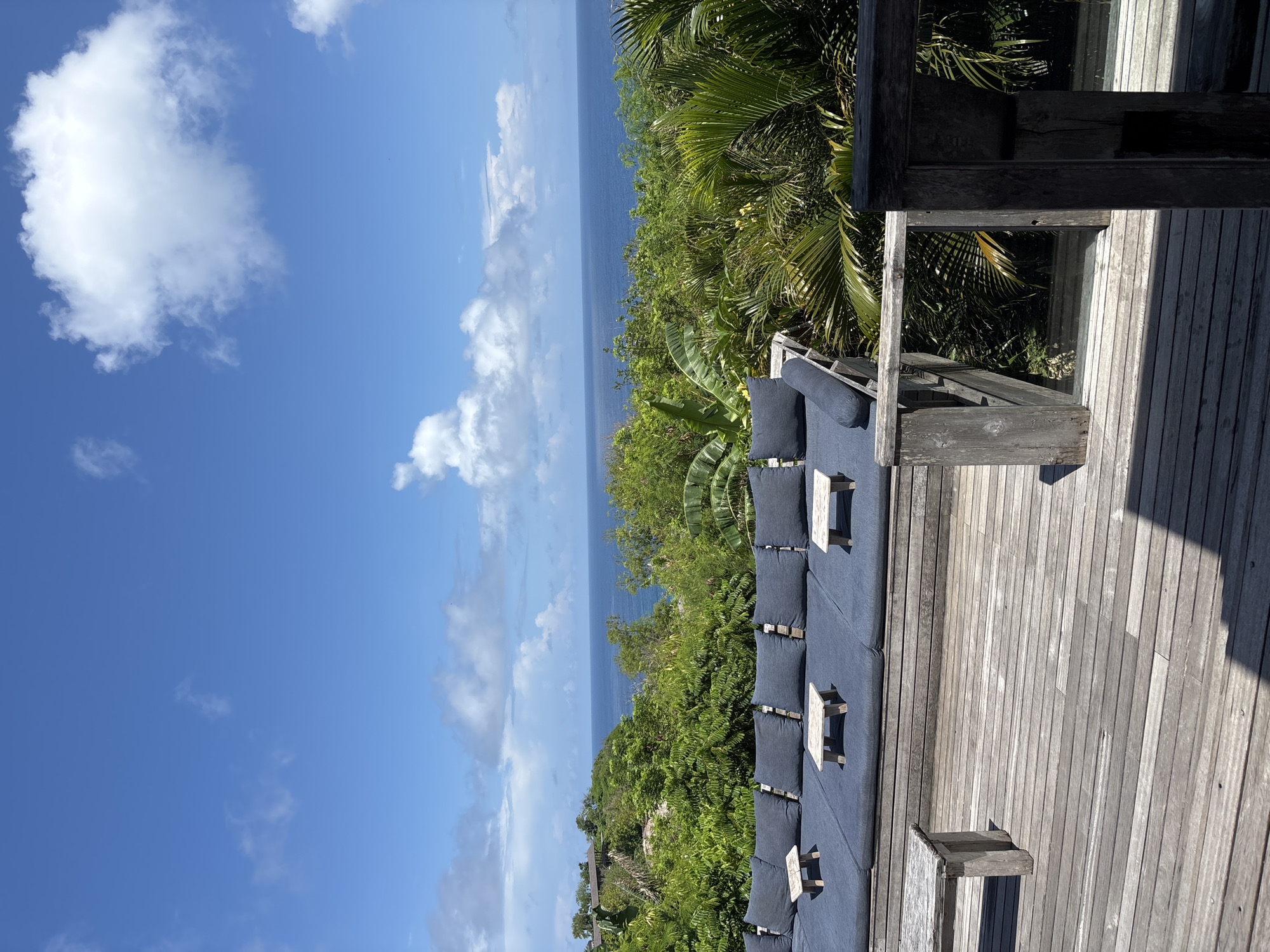 The image depicts an outdoor seating area likely in Uluwatu, Indonesia, characterized by a wooden deck furnished with long benches covered in navy cushions and paired with small, white, wooden tables. Lush, tropical vegetation, including palm trees and other green foliage, surrounds the deck, creating a backdrop towards the horizon. The blue ocean is visible in the distance, stretching to the horizon line under a partly cloudy sky. The sky is a clear blue with scattered white clouds. The scene is viewed from an elevated angle, providing a wide perspective of the seating area, the surrounding vegetation, and the distant ocean. A wooden structure, possibly part of a building or shaded area, is positioned on the right side of the frame, casting a shadow onto the deck.