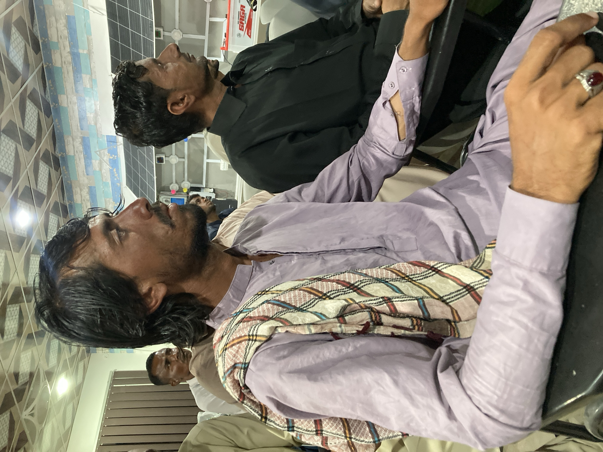 The image shows two men sitting in chairs. The man in the foreground is wearing a light purple shirt and a scarf. The man in the background is wearing a black shirt. They appear to be looking up at something. The environment appears to be indoors, potentially an office setting, given the lighting and the partial view of what appears to be a commercial fixture in the background. There are other people in the background as well, though they are out of focus.