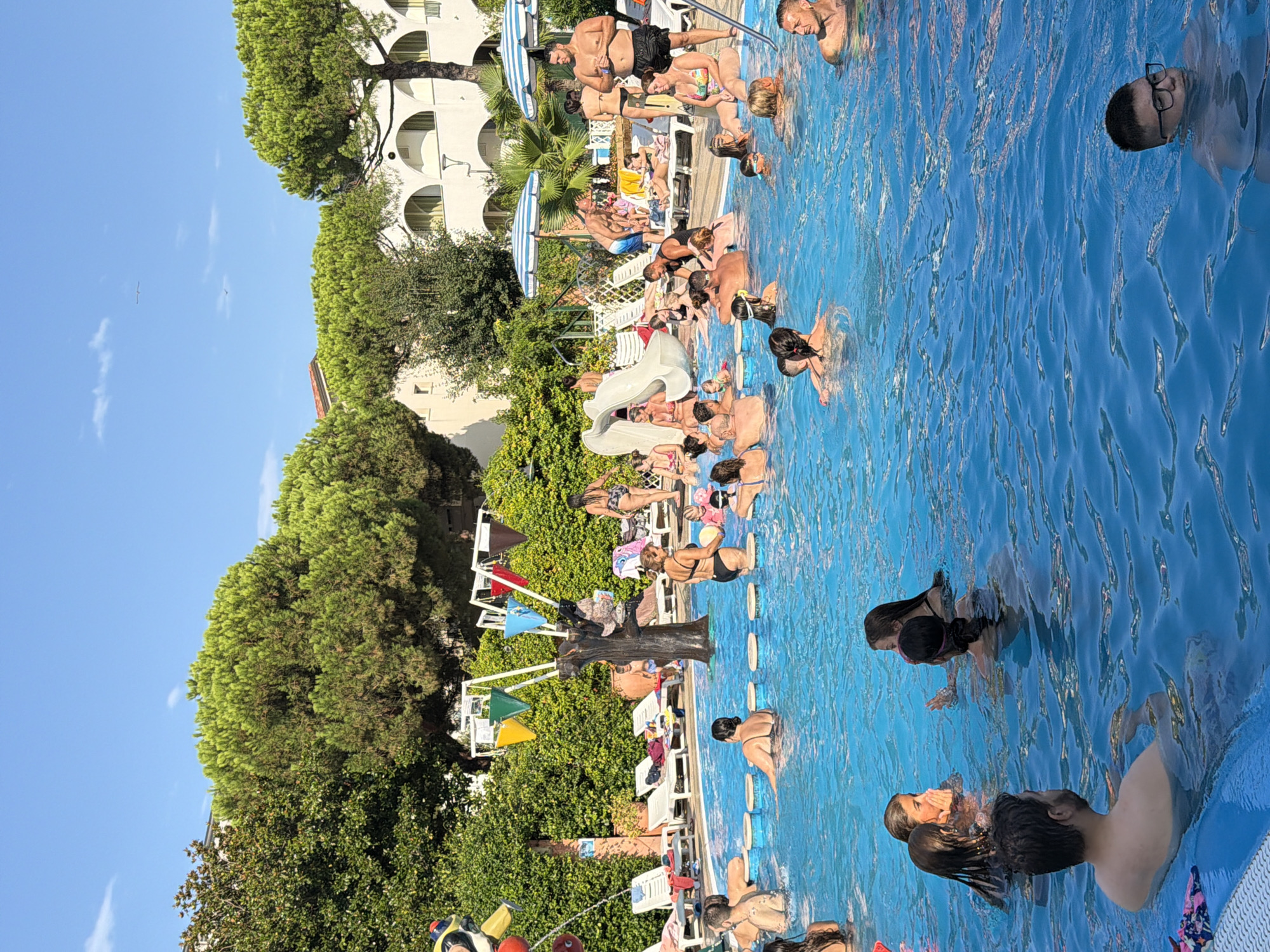The image captures a busy outdoor swimming pool scene under a clear blue sky. Numerous people are engaged in various activities within the vibrant blue water. In the foreground, a man with short hair and glasses is partially submerged. To his left, another man is swimming. In the middle ground, a group of four young women are interacting in the water, with two of them appearing to be whispering. To the right, a man with a muscular build is standing and looking towards the left. A woman in a colorful swimsuit is sitting on the edge of the pool, holding a yellow ball. Further back, a water slide is visible, and several individuals are lounging on white chairs surrounding the pool, some with umbrellas providing shade. The background features lush green trees and a light-colored building with arched windows. The scene appears to be a recreational area, possibly a hotel or resort, in Gatteo, Italy.