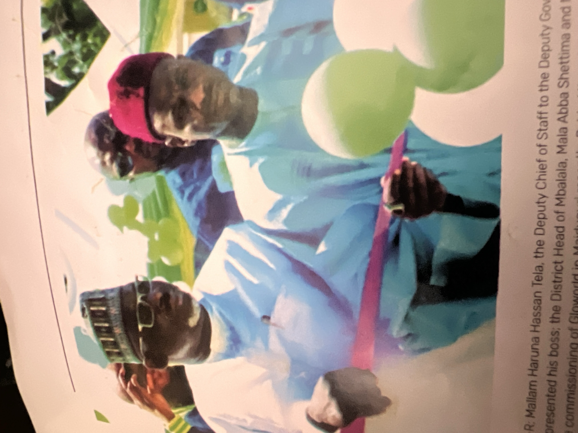The image shows two men in traditional attire, likely at an outdoor event. The man on the left, wearing a blue and white patterned cap and glasses, appears to be cutting a pink ribbon with scissors. He is dressed in a white, long-sleeved garment. To his right, a man in a red fez-style hat and a light blue garment looks towards the camera. In the background, other individuals are visible, some wearing blue shirts. Several green balloons are in the foreground. The text at the bottom of the image mentions "Mallam Haruna Hassan Tela, the Deputy Chief of Staff to the Deputy Governor" and the "commissioning of Glowworld in Maiduguri." While the text mentions Maiduguri, the question specifies Jalingo, Nigeria. Without further context or visual cues linking the image to Jalingo, it's impossible to confirm that location. The scene suggests a formal event, possibly a business or public ceremony, given the ribbon-cutting and balloons. The lighting suggests it is daytime.