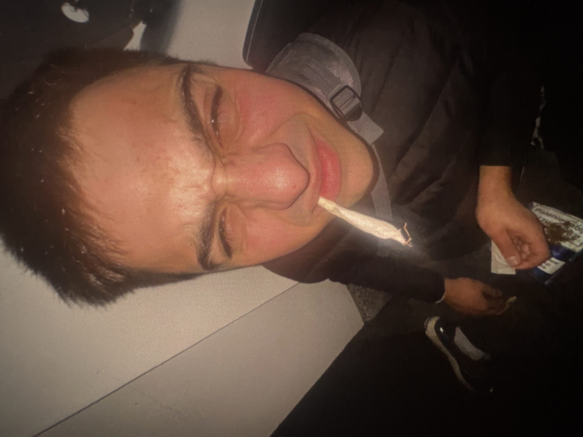A close-up, low-angle, distorted shot shows a young man with dark, short hair, fair skin, and brown eyes. He is squinting and making a grimacing face, with his eyebrows furrowed and cheeks flushed. A hand-rolled cigarette is held between his lips. He is wearing a dark jacket with a gray collar and strap. In his right hand, he holds a small, folded packet, possibly containing tobacco or other materials, and a small portion of what appears to be cannabis is visible. The background is dark and out of focus, with a textured wall on the left side and a dark surface on the right, possibly indicating an indoor setting. No specific location in Warsaw, Poland is discernible from the image.