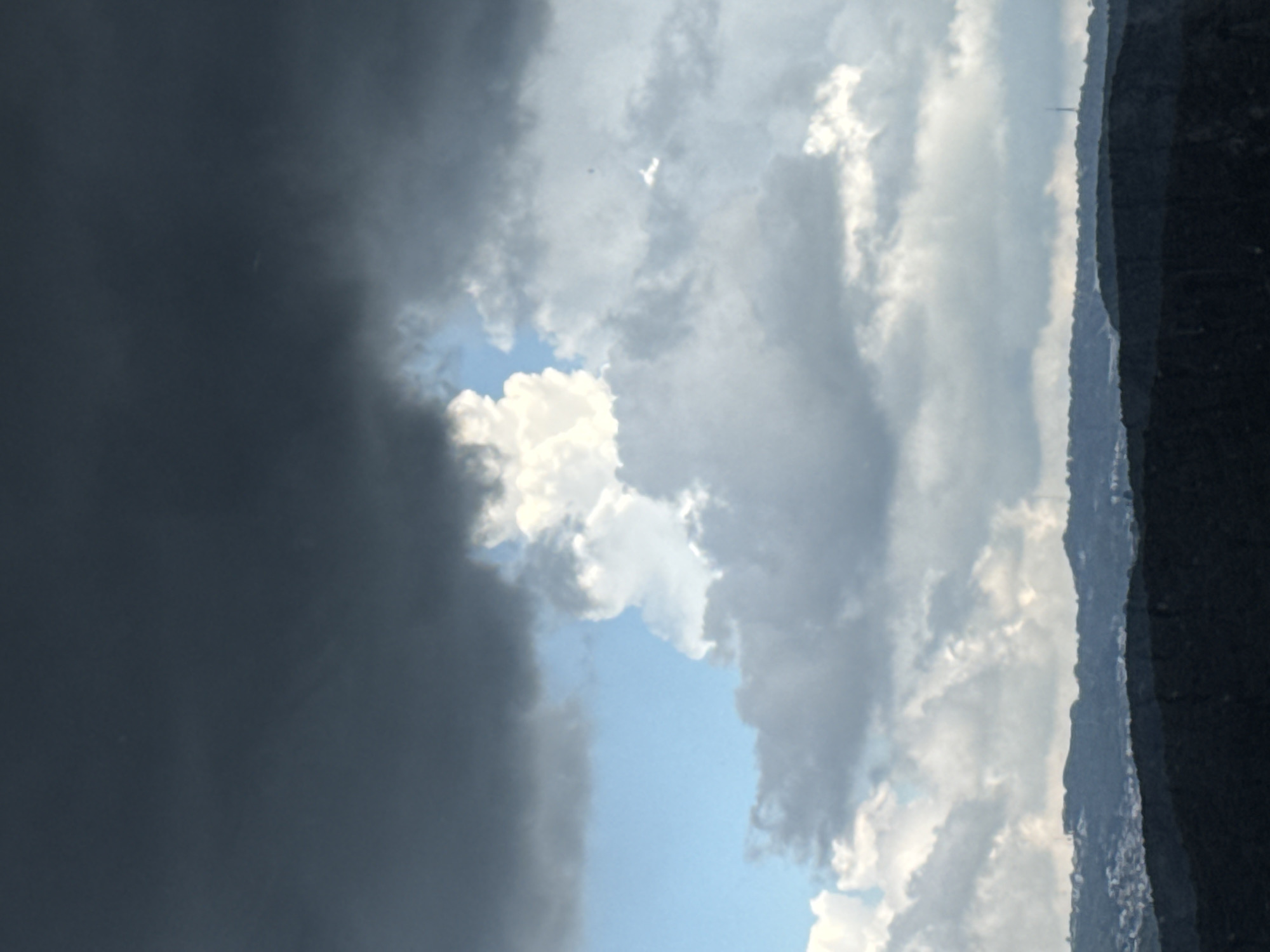 The image captures a dramatic sky over a hilly landscape, likely in or near Đa Lộc, Vietnam. The upper portion of the frame is dominated by dark, heavy storm clouds, indicative of impending rain or a passing tempest. Below these ominous clouds, patches of bright blue sky peek through, interspersed with lighter, fluffier clouds. The contrast between the dark and light clouds creates a sense of depth and atmospheric movement. The horizon is marked by a series of dark, undulating hills that recede into the distance. The foreground hills are densely covered in vegetation, appearing almost black in the low light. A scattering of buildings is faintly visible on the slopes of the middle hills, suggesting human habitation. A tall, slender structure, possibly a communication tower, is discernible on the furthest ridge. The overall impression is one of nature's power and vastness, with a hint of human presence in the rugged terrain.