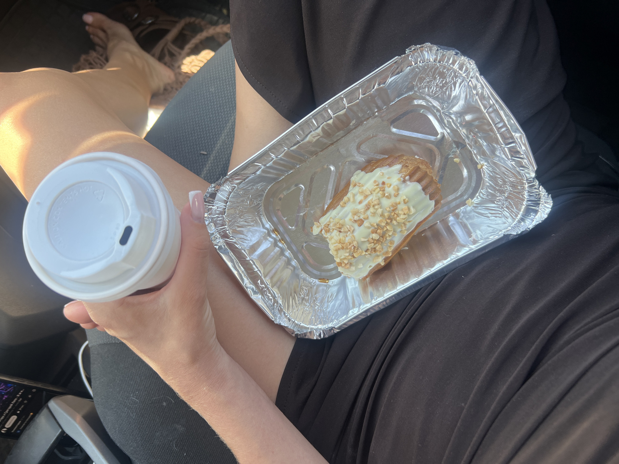 This is a first-person perspective photograph, likely taken inside a vehicle, showing a person holding a white disposable coffee cup in their left hand and an eclair in a silver foil container in their right hand. The person is wearing a dark brown dress. Sunlight is casting shadows and highlighting the person's legs and arms, suggesting it is daytime. The background shows a car seat and a hint of a woven bag, possibly indicating a travel or outing context. The eclair appears to be topped with white icing and chopped nuts. Visible text on the coffee cup lid is partially obscured but appears to be a warning symbol and some text. The image has a casual and relaxed feel, possibly capturing a moment of enjoying a snack and drink. Given the location context of Budva, Montenegro, it's plausible this is a scenic stop during a trip.