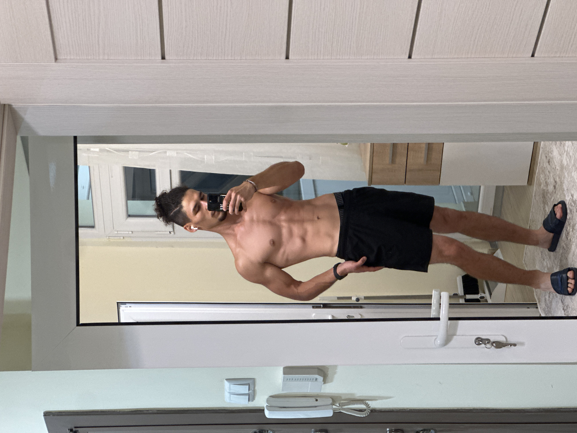 A young man with dark hair styled in a bun and a beard is taking a mirror selfie in an indoor setting. He is wearing black shorts and is shirtless, showcasing his toned physique. He has earbuds in his ears and a bracelet on his wrist. The environment appears to be a hallway or entryway, with a white door on the right and a door with a handle and lock on the left. A wall-mounted intercom system is visible on the left wall. In the background, a dresser and a window with sheer curtains can be seen. The floor is covered with a rug. The scene is lit by indoor lighting. No specific location in Varna, Bulgaria is identifiable.