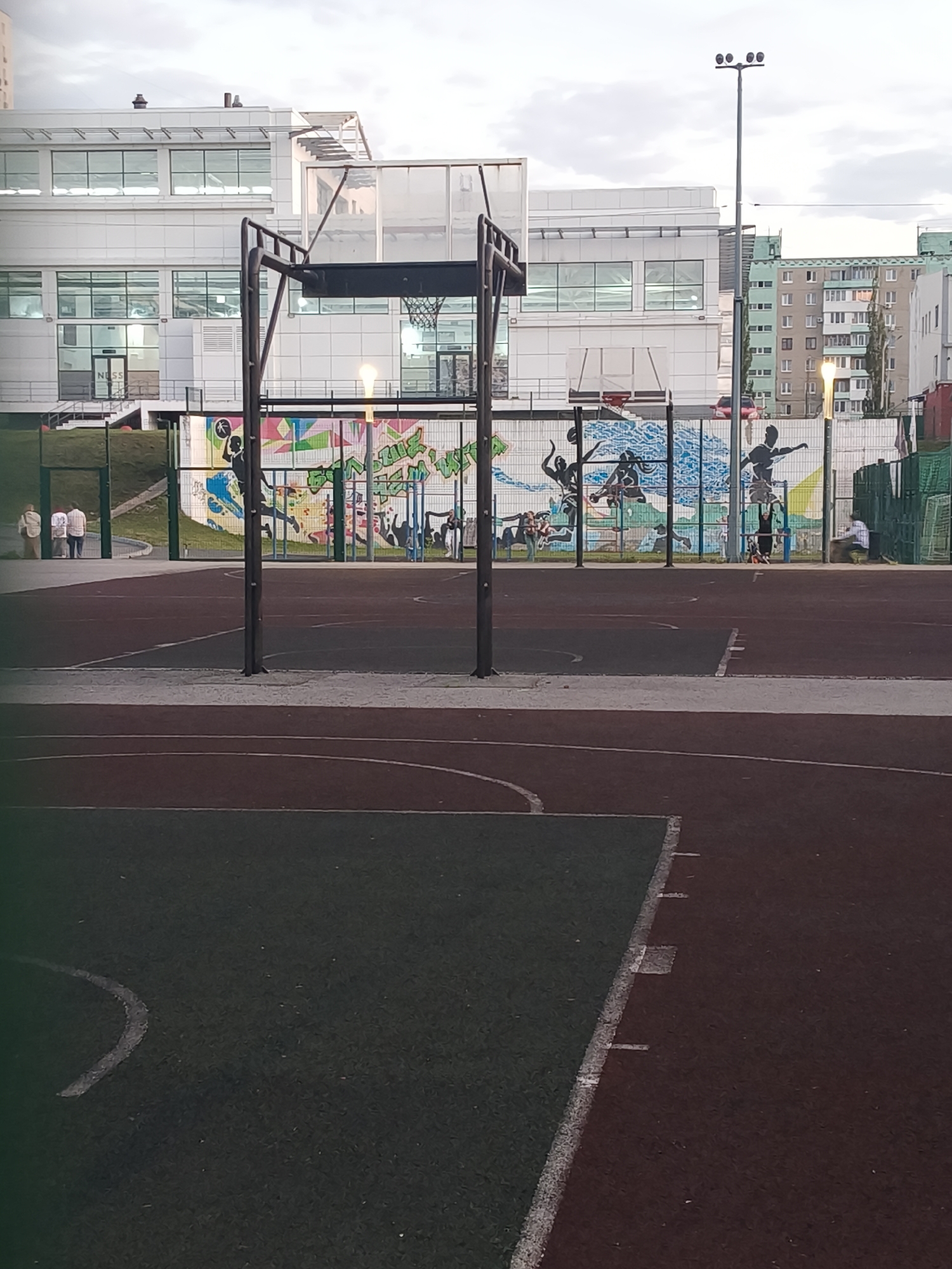 An outdoor basketball court is shown in the foreground, with a dark red surface and green sections marked with white lines. In the background, a white building with multiple windows and a glass facade is visible. Behind the building, a residential building with several floors and windows is depicted. A tall lamppost stands in the middle of the scene, and a mural with colorful graffiti and silhouettes of people playing sports adorns a fence. A few individuals are present on the court and near the fence. The sky is overcast with clouds.