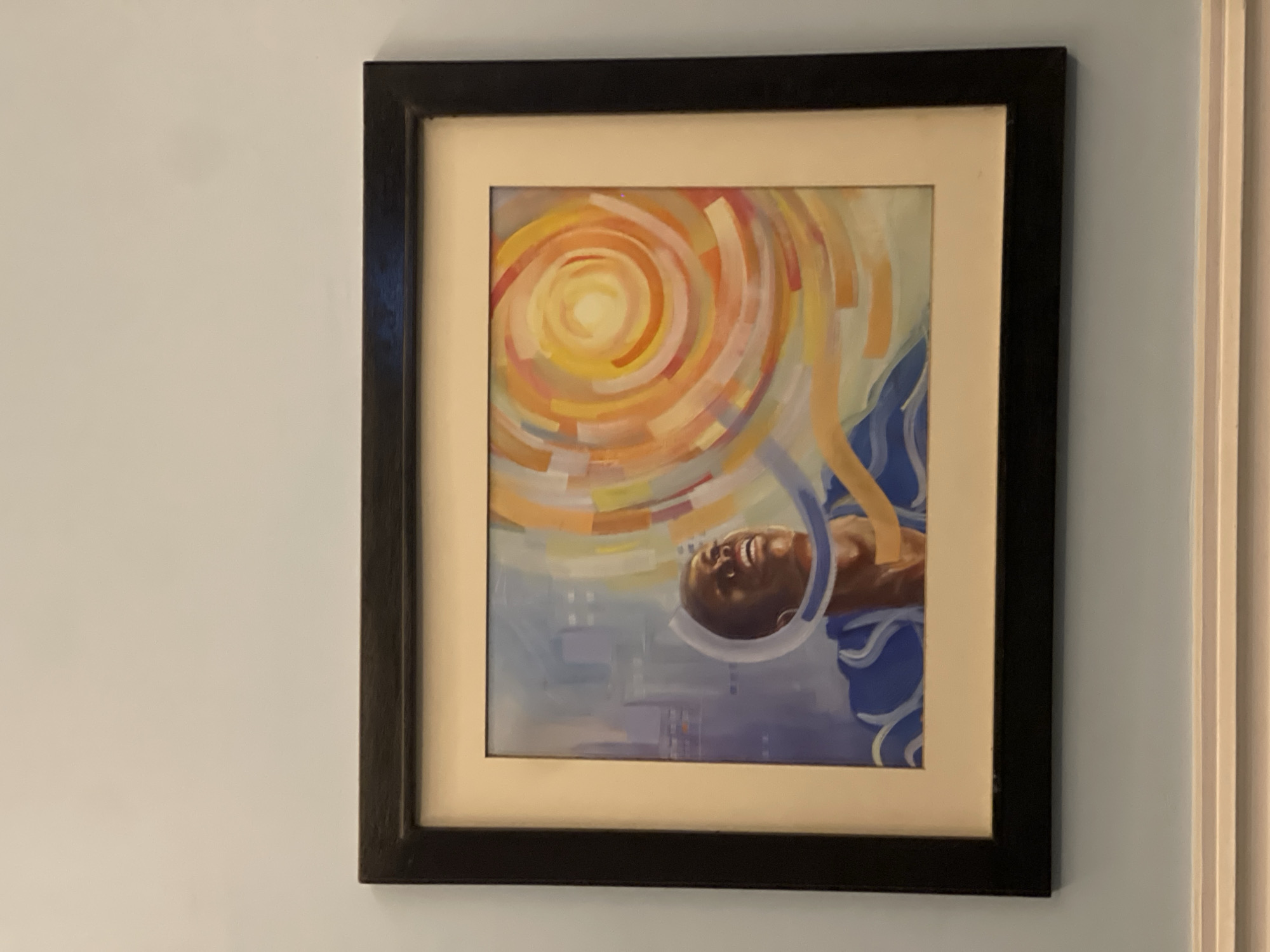 A framed painting hangs on a light blue wall next to a white door frame. The painting depicts a man with dark skin, possibly of African descent, surrounded by a yellow and orange spiral design that evokes a sun-like aura. The man appears to be looking upwards and smiling. He has a white halo around his head. The background contains bluish and white hues. The painting is framed with a black frame and a neutral-colored mat. There is no indication of the location in the city of Jos, Nigeria.