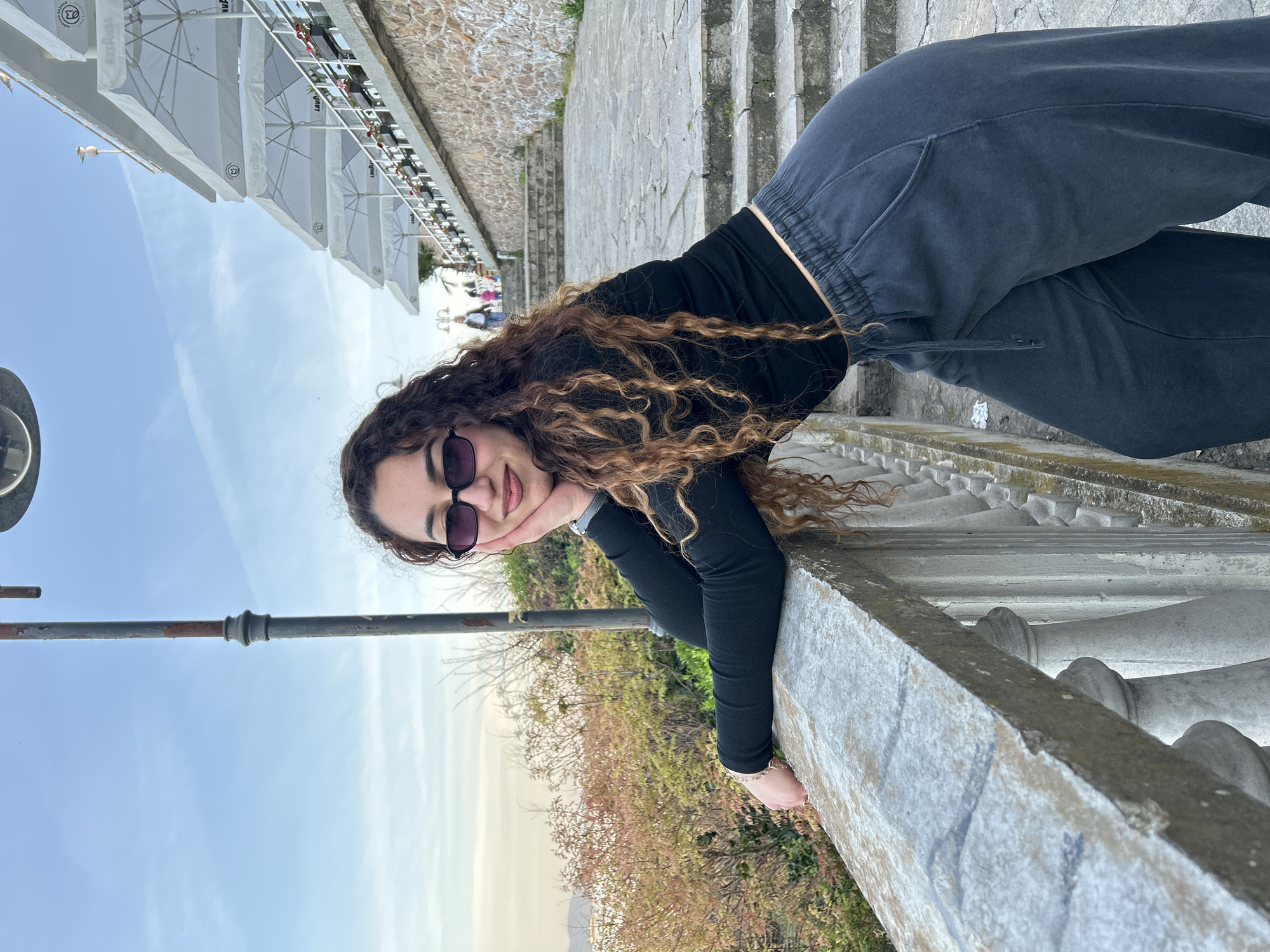 An outdoor, daytime portrait features a young woman with long, curly brown hair, wearing dark sunglasses, a black long-sleeved top, and dark grey sweatpants. She leans casually on a weathered stone balustrade, smiling gently with her chin resting on her left hand. A delicate bracelet adorns her left wrist.
The image is oriented on its side, showing a clear, light blue sky with a few faint clouds dominating the upper portion. In the background, behind the woman, a series of stone steps ascend alongside a textured stone wall. To her left, a sturdy metal railing extends into the foreground.
Beyond the balustrade and a line of trees and bushes exhibiting both green and brownish-red foliage, hints of a distant landscape are visible. Further in the background, several white outdoor awnings or large umbrellas are arranged in a row, suggesting a cafe or restaurant area. Faint text, including "LAVAZZA" and possibly "MERCIA" or "MERCI" along with a circular logo, can be seen on these awnings. A few other indistinct figures are visible near the awnings. The setting appears to be an elevated viewpoint or promenade in Burgas, Bulgaria, on a clear, pleasant day.