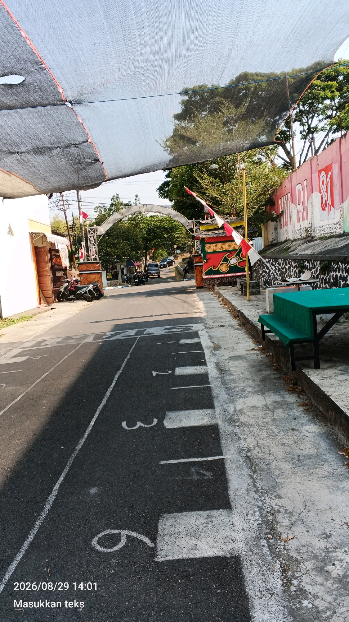 The image captures a street scene in Mulyorejo, Indonesia, under a large, dark mesh sunshade. The road surface is marked with white lines and numbers, suggesting a designated area. On the right side of the road, a green picnic table and benches are visible, accompanied by a low stone wall. Further down the street, an arched gateway stands, decorated with flags and colorful banners. Several motorcycles are parked near buildings on the left side of the road, and a few cars are seen in the distance. The environment appears to be a street in a developed area, with buildings, trees, and infrastructure visible. The time stamp "2026/08/29 14:01" and the text "Masukkan teks" (Insert text) are overlaid on the bottom left corner of the image.