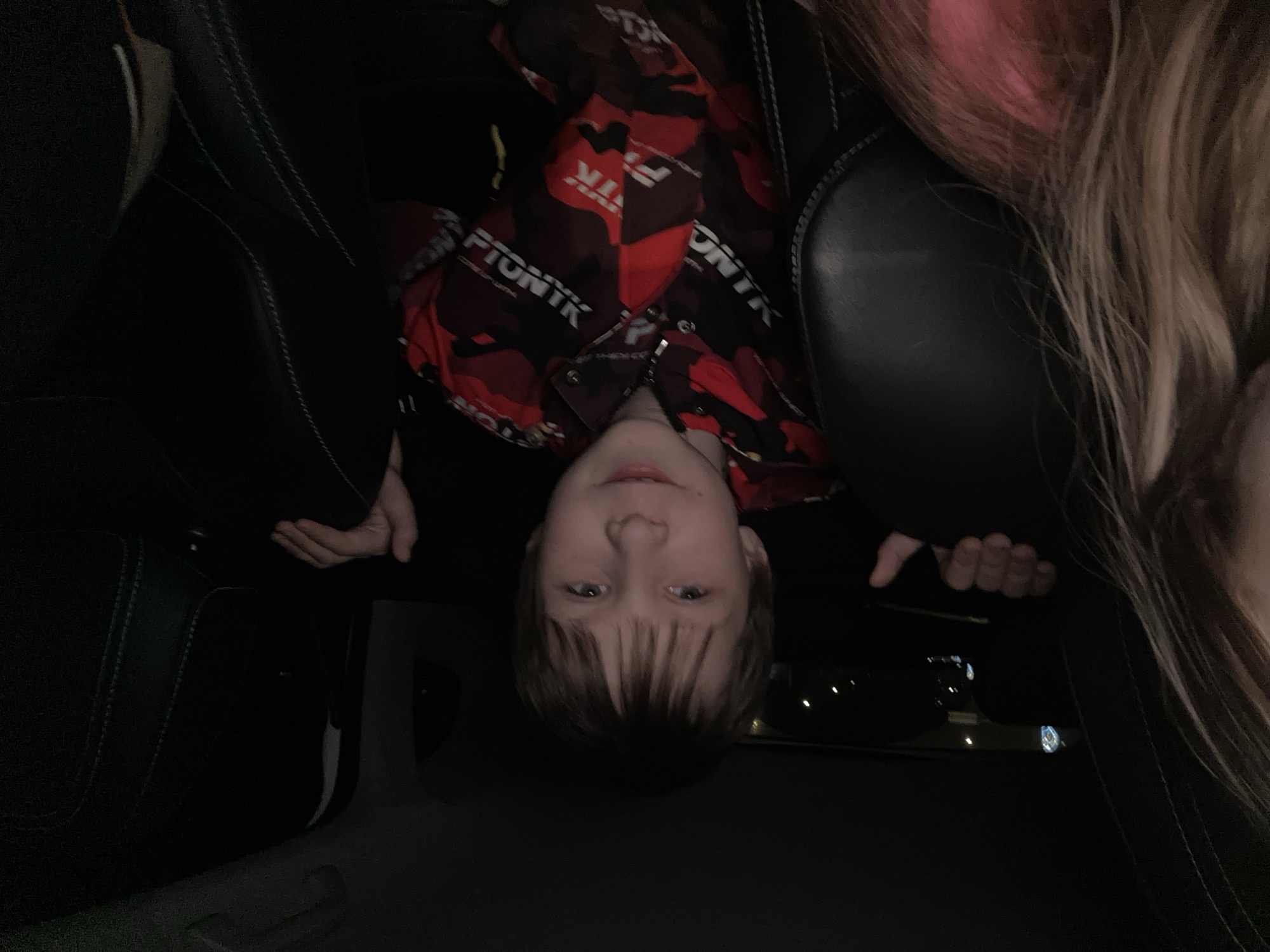 A child is seen upside down in the back seat of a car. The car's interior is dark, and the city lights are visible through the window. The child is wearing a red and black camouflage jacket. Another person with long blonde hair is partially visible in the scene. No data is available about Moscow, Russia.