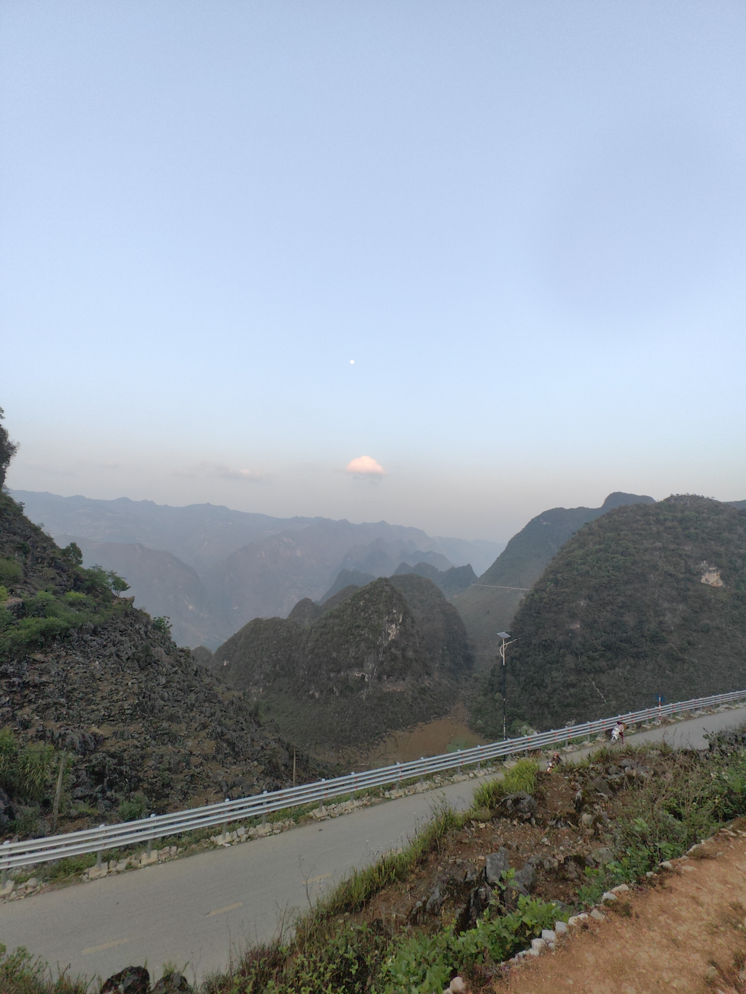 The image depicts a mountainous landscape in Đồng Văn Commune, Vietnam. The foreground features a paved road with a metal guardrail, bordered by rocky terrain and vegetation. In the midground, a series of green, jagged mountains rise, partially obscured by haze. A white pole structure with a solar panel is visible beside the road. A motorcycle with two people is on the road. In the background, more mountains fade into the distance under a pale sky with a thin cloud. The moon is also visible in the sky.