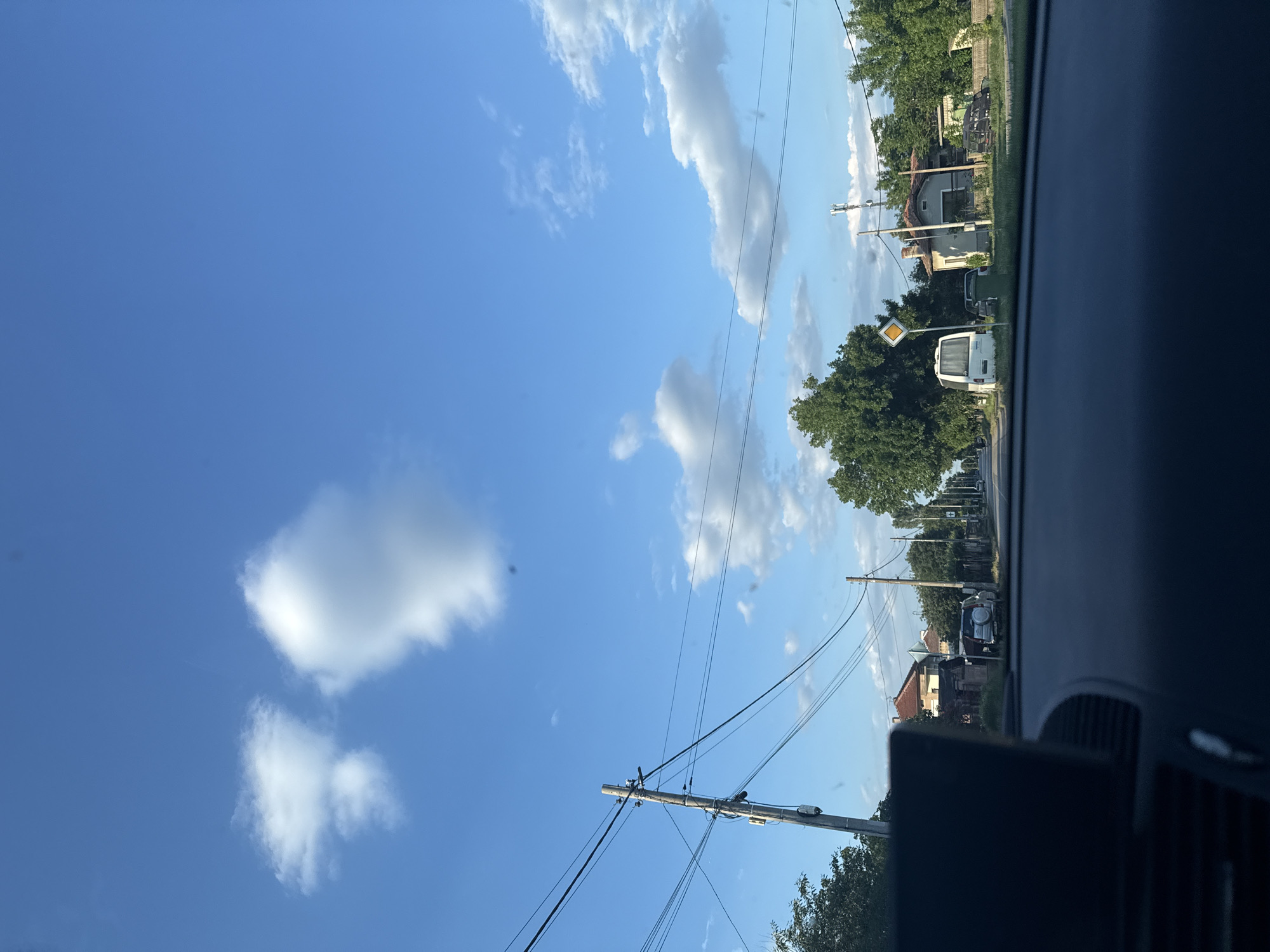 The image depicts a street scene in what appears to be a residential area, possibly within or near Sarnevo, Bulgaria. The view is from inside a vehicle, likely a car, looking out through the windshield. The sky is blue with scattered cumulus clouds. Power lines stretch across the sky.
On the right side of the road, there are houses and trees lining the street. A yield sign is visible on the roadside. A white van is parked further down the street. There are a few other cars visible in the distance. The interior of the vehicle, specifically the dashboard and window frame, are visible in the foreground.