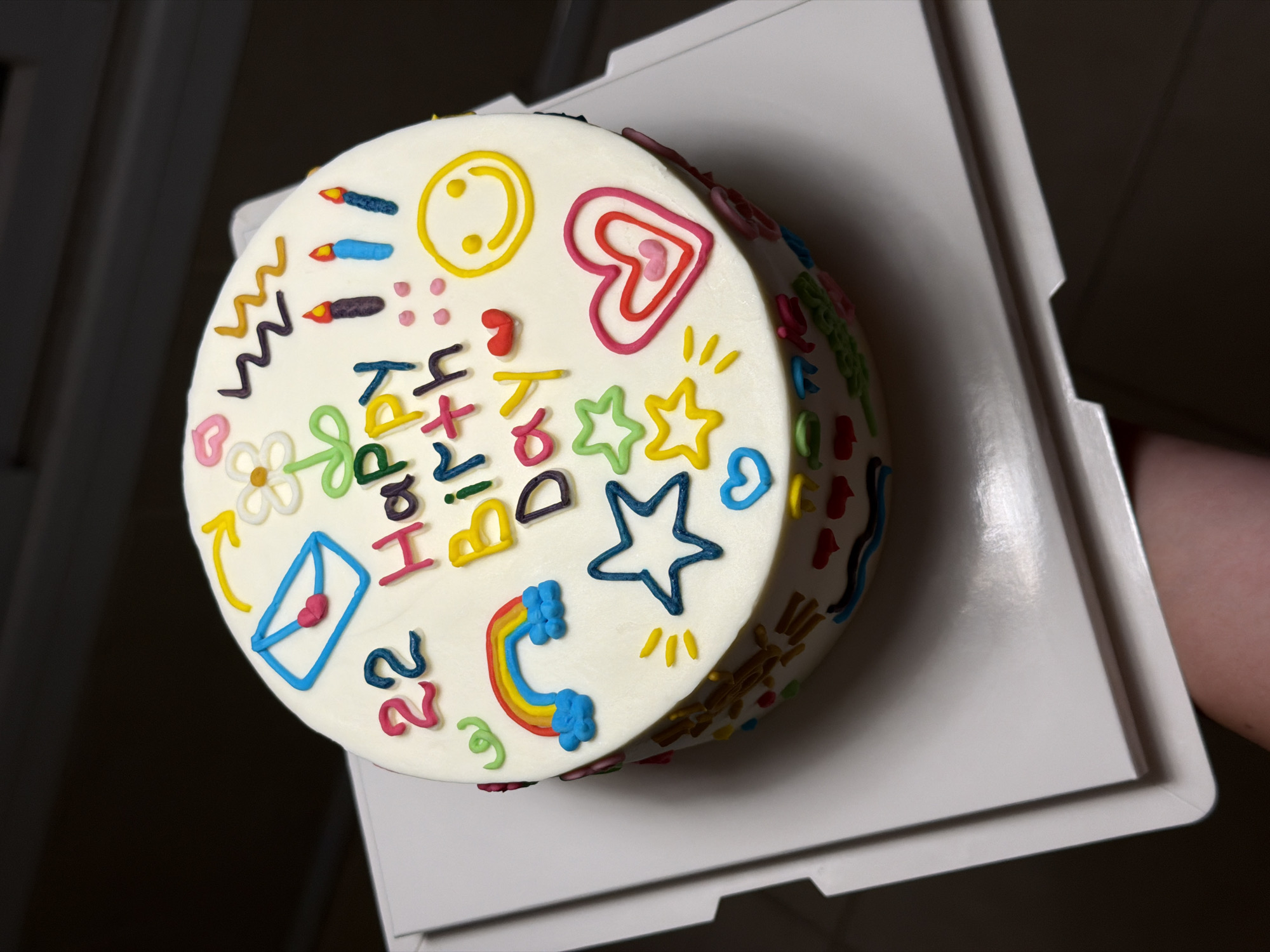 A round, white frosted cake decorated with colorful icing is presented on a white cake stand. The cake is adorned with various playful designs, including the words "22 Happy Birthday" written in vibrant icing. Also visible are a blue envelope with a pink heart, a yellow smiley face, a rainbow with clouds, stars, candles, and a red heart. The side of the cake is decorated with a medley of small, colorful shapes. The cake is held by a hand, partially visible at the bottom right of the frame, suggesting it is being presented or served. The background is dark and out of focus, with a hint of a wall or furniture. There is no discernible information within the image to suggest a specific location in Tel-Aviv, Israel.