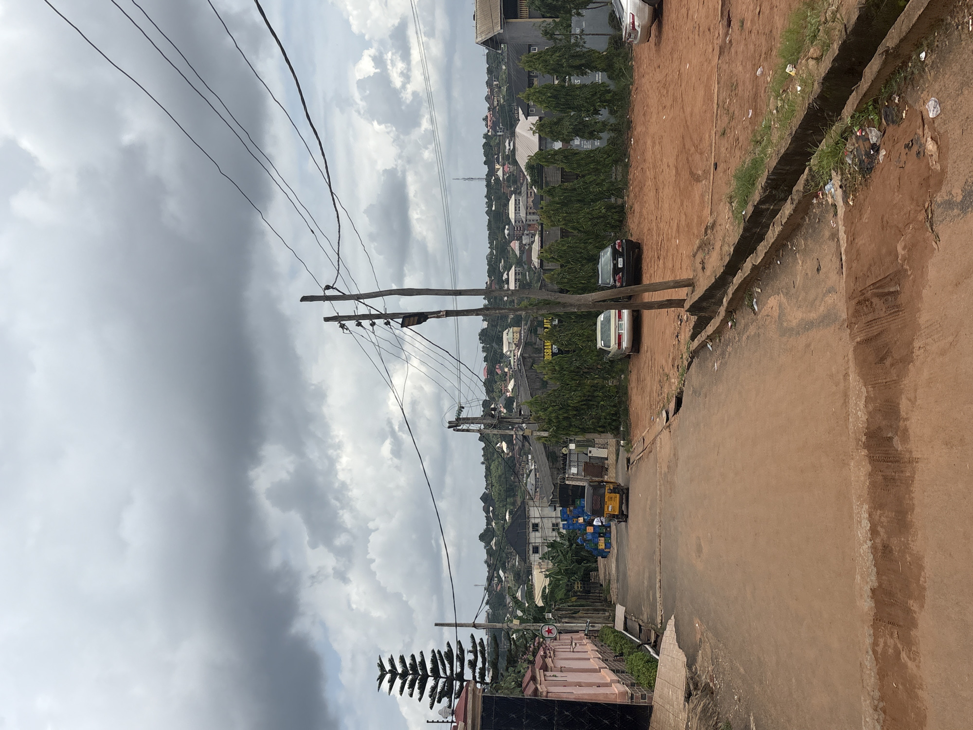 The image captures a view of a street in Nkpor, Nigeria, under a cloudy sky. The foreground shows a reddish-brown dirt road with some patchy water or mud. To the left, a light pink building with black accents and a tall, conical evergreen tree are visible. Further down the road, a yellow auto-rickshaw is being driven by an unseen individual. On the right side of the road, a drainage ditch is filled with debris, including plastic bags and other trash. Beyond the ditch, an open area of red dirt is present, with several parked cars, including a silver sedan and a black sedan. In the background, a densely populated urban landscape with numerous buildings of varying sizes and architectural styles stretches towards the horizon, interspersed with greenery and trees. Overhead, power lines crisscross the sky, supported by wooden utility poles. The overall scene depicts a typical street in a developing urban area, characterized by a mix of residential and commercial structures, unpaved or partially paved roads, and the presence of everyday vehicles.