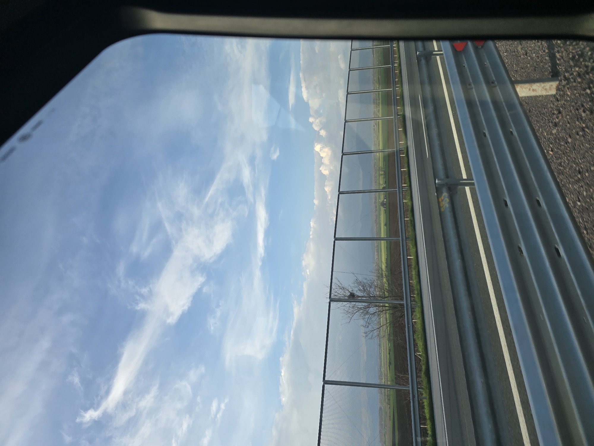 The image appears to be taken from inside a vehicle, looking out a side window. A bright, blue sky with wispy white clouds is visible. A sound barrier, constructed of vertical supports with translucent panels between them, runs along the right side of the frame. Beyond the sound barrier is a field with some sparse vegetation. A highway is visible to the right of the sound barrier, with multiple lanes marked by white lines. The asphalt surface of the highway appears to be dark in color. The image provides insufficient data to determine if it is in the city of Chokoba, Bulgaria.