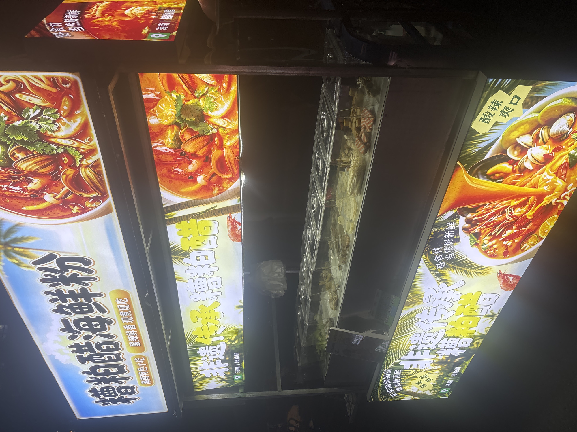 This is a nighttime photograph of a food stall in Suzhou Industrial Park, China, likely a street food vendor. The stall has multiple illuminated signs and displays showcasing food items.
The uppermost sign features large Chinese characters that translate to "Claypot Seafood Noodles," accompanied by smaller text indicating "Hainan Specialty Snack" and "Sour, Spicy, Fragrant, Freshly Made." A vibrant image of a seafood soup or stew with noodles, shrimp, and clams is prominently displayed on this sign.
Below this, another illuminated sign repeats "Claypot Seafood Noodles" in large characters, along with "Non-heritage Inheritance." This sign also features a tropical background with palm trees and an image of a seafood dish.
The stall itself has a display case with various ingredients for the noodles, including what appears to be vegetables, seafood, and possibly tofu or processed meat. Some of these ingredients are in clear plastic containers. Prices are visible on a small sign in front of the display case.
The overall environment suggests an outdoor food market or street-side operation in an urban setting. The lighting is artificial, creating a bright and eye-catching presentation for the food. There are no people clearly visible in the frame.