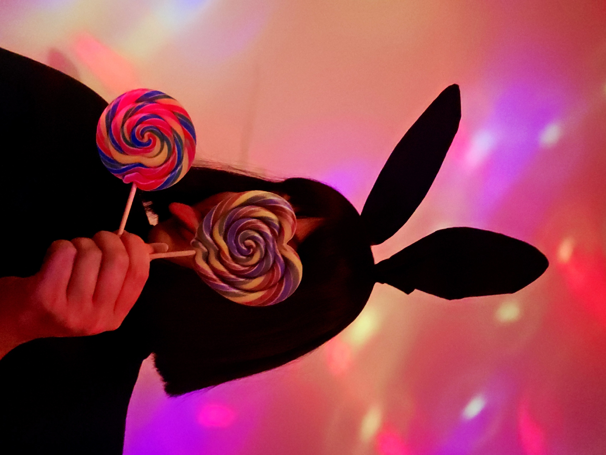 A person wearing bunny ears holds two lollipops. The background has an unknown origin and is brightly illuminated by red, pink, and purple lights. The person is wearing a dark-colored shirt. The lollipops have a swirl design in various colors. The person's skin appears red from the lighting. The image's location is unknown.