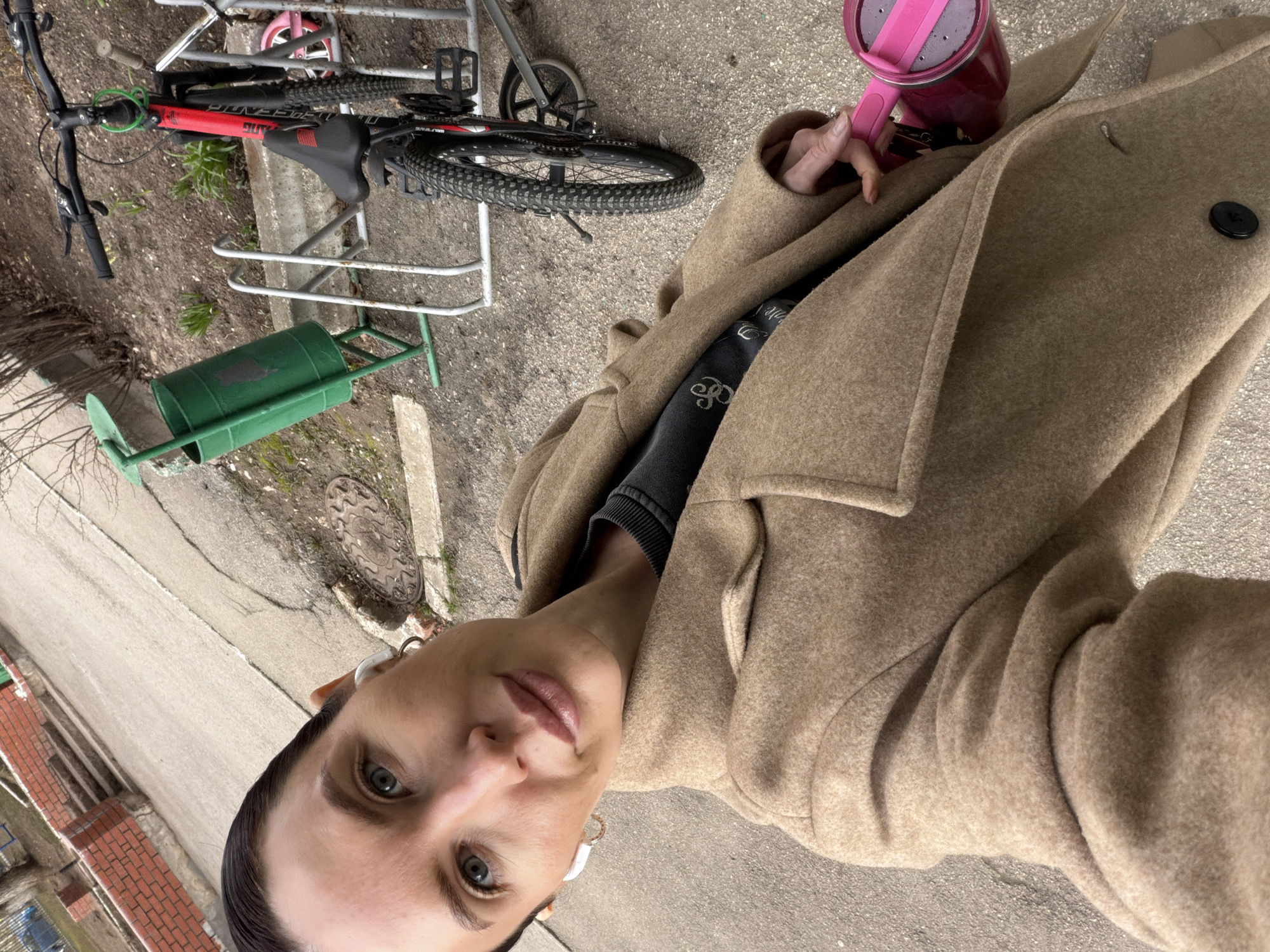 A selfie-style portrait captures a woman outdoors in Perm, Russia, with the image rotated 90 degrees clockwise. The woman, positioned on the right side of the frame, has dark hair pulled back, light-colored eyes, and a neutral expression with light-colored lipstick. She wears small white earbuds in both ears and a subtle hoop earring in her right ear.
She is dressed in a warm, light brown/tan long coat featuring a wide lapel and a black button closure. Underneath, a dark grey or black sweater is visible, bearing a partial white logo that appears to be "BALENCIAGA" (parts like "BALEN" and "AGA" are discernible). In her right hand, held near her chest and partially obscured by her coat, is a bright pink and purple travel mug or portable blender.
The background depicts an urban or suburban outdoor setting. A red and black mountain bike, with the brand "GIANT" visible on its frame, is parked on a metal bicycle rack. Behind it, a small pink bicycle wheel is also present. On the ground next to the bike rack, there is a distinct green, barrel-shaped object with handles, possibly a planter or a decorative item. The ground is paved, likely asphalt or concrete, and a concrete curb runs along the scene. A round, ornate metal manhole cover is visible on the pavement. Sparse, dry branches and minimal vegetation are scattered, suggesting either a cooler season or dormant plant life. In the distance to the left, a brick wall structure can be seen. The lighting indicates it is daytime, possibly a cool or overcast day given the woman's warm attire.