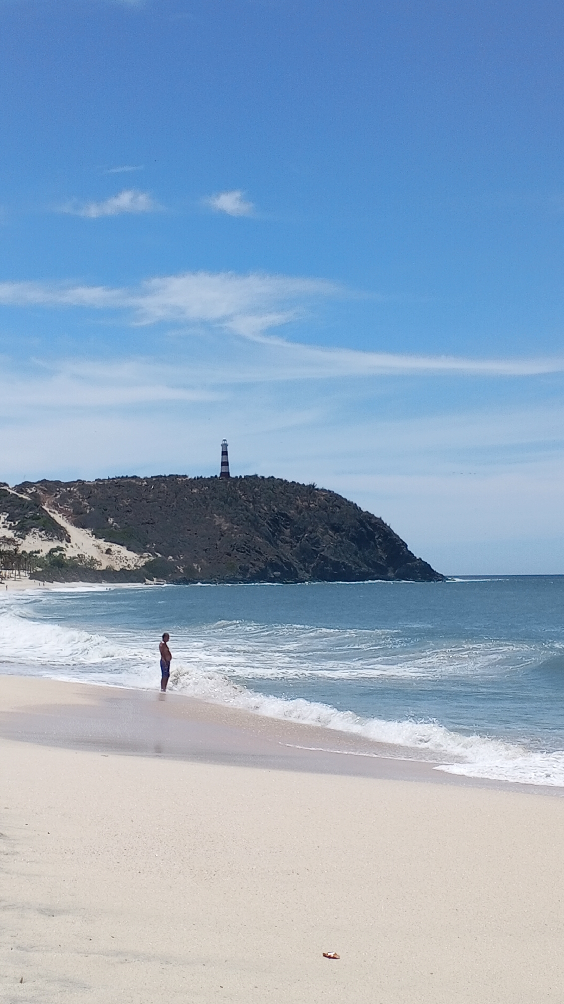 The image depicts a beach scene on a sunny day. A person stands near the water's edge, facing the ocean. In the background, a hill rises with a lighthouse on top. The sky is blue with scattered clouds. The sand is light-colored and the water is blue-green with visible waves. An orange object is visible on the sand. Location is Unknown, Unknown.