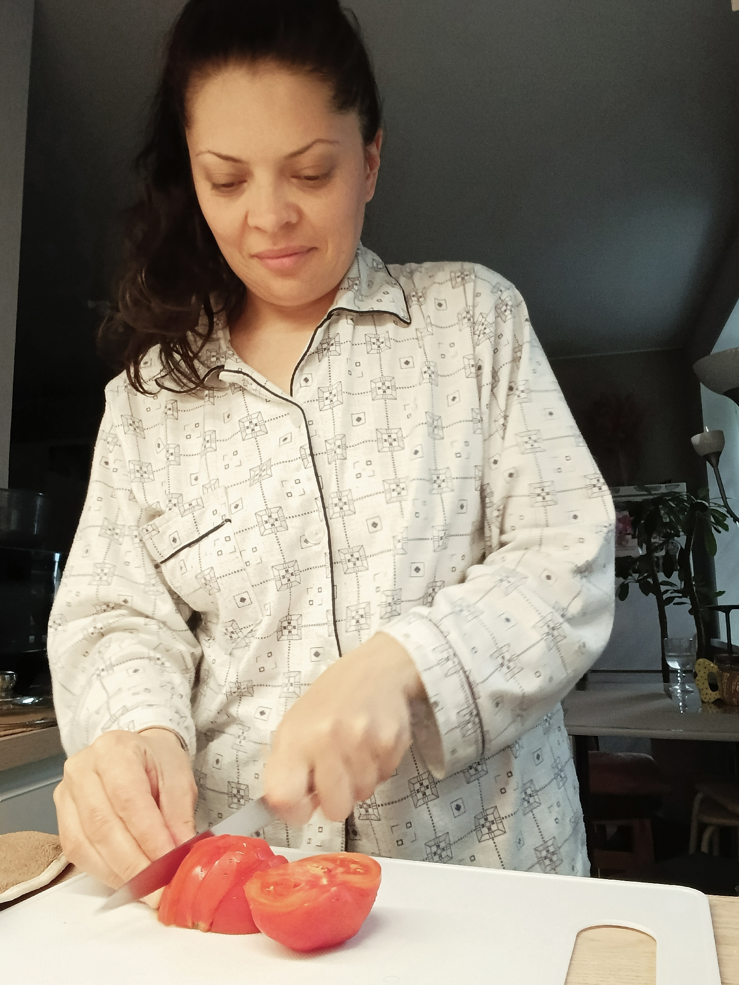 A woman with dark hair pulled back is standing in a kitchen, wearing a light-colored pajama top with a geometric pattern. She is looking down and slicing a tomato on a white cutting board with a silver knife. The background is dimly lit and shows a kitchen counter with some objects, including a plant and glassware. The scene appears to be indoors. The location is not specified in Saratov, Russia.