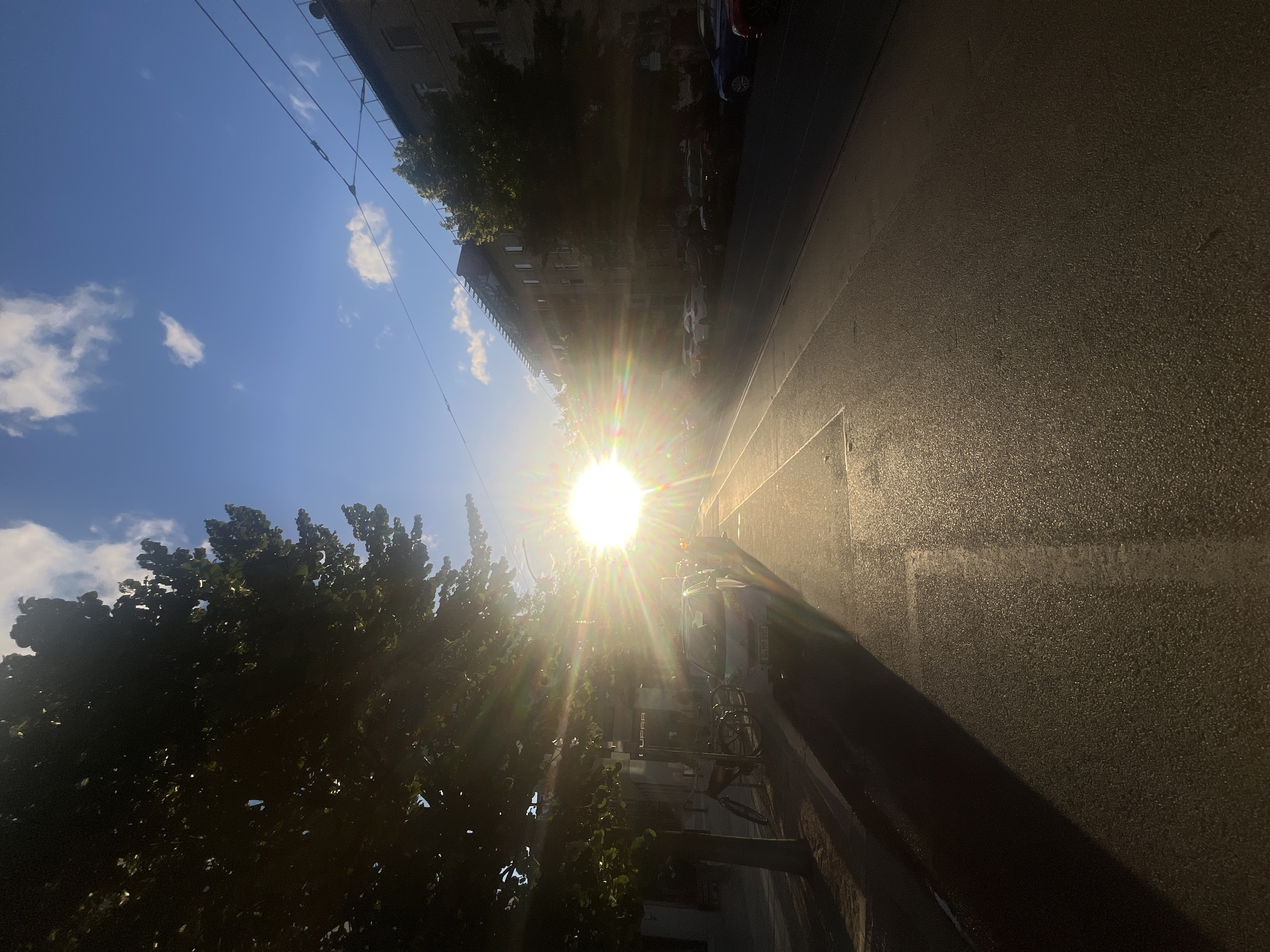 The image depicts a street scene in Berlin, Germany, during what appears to be late afternoon or early evening, given the low angle of the sun. The sun is prominently featured, casting a bright glare and lens flare across the frame, situated just above the center of the image. The sun's rays illuminate the wet asphalt of the street, creating a shimmering effect.
On the left side of the street, a tree with dense foliage partially obstructs the view. In front of the tree, a small white car is parked, and further back, several bicycles are visible. The sidewalk area shows some storefronts, one of which has signage that appears to read "CUPRA".
The street itself has tram tracks running along its length. On the right side of the street, buildings line the road, with trees in front of them. Several cars are parked along the curb on this side as well. The sky is blue with scattered white clouds. The overall impression is one of a typical urban environment bathed in the warm, low light of the setting sun.