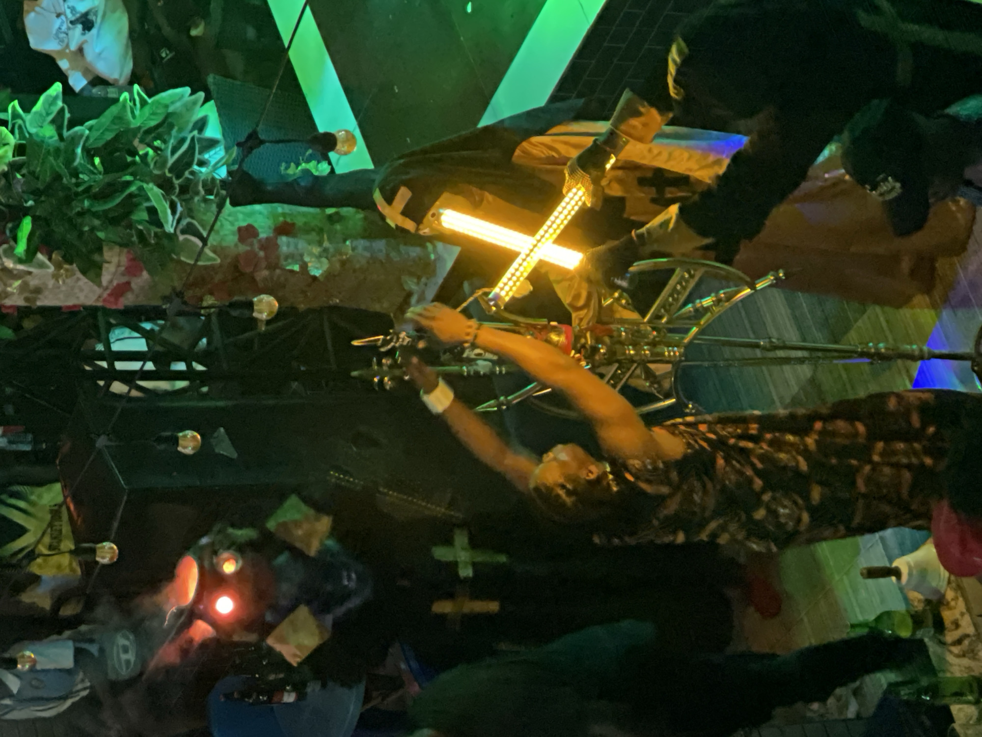 The image captures a dimly lit, indoor scene with several individuals in costumes or elaborate attire. In the foreground, a person in a patterned dress is interacting with a metallic structure adorned with lights. To their right, another individual wears a dark hooded robe over a yellow garment with a prominent cross emblem, holding a glowing, cross-shaped light fixture. Behind them, a costumed figure with red glowing eyes and a cauldron-like headpiece stands, emitting smoke. The environment appears to be decorated with artificial plants and hanging lights. In the background, glimpses of bottles and other items suggest a bar or event space. The overall atmosphere is theatrical and slightly eerie, with vibrant colored lighting.