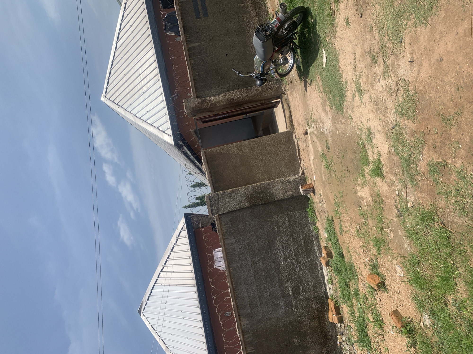 This image depicts an outdoor scene in Jauro Yinu, Nigeria, featuring two residential buildings with corrugated metal roofs. A grey concrete wall, topped with barbed wire, encloses the property. To the right, an open doorway leads into one of the buildings. A motorcycle is parked on the dirt ground to the right of the entrance. Scattered bricks are visible on the ground in the foreground and near the wall. The sky is partly cloudy.