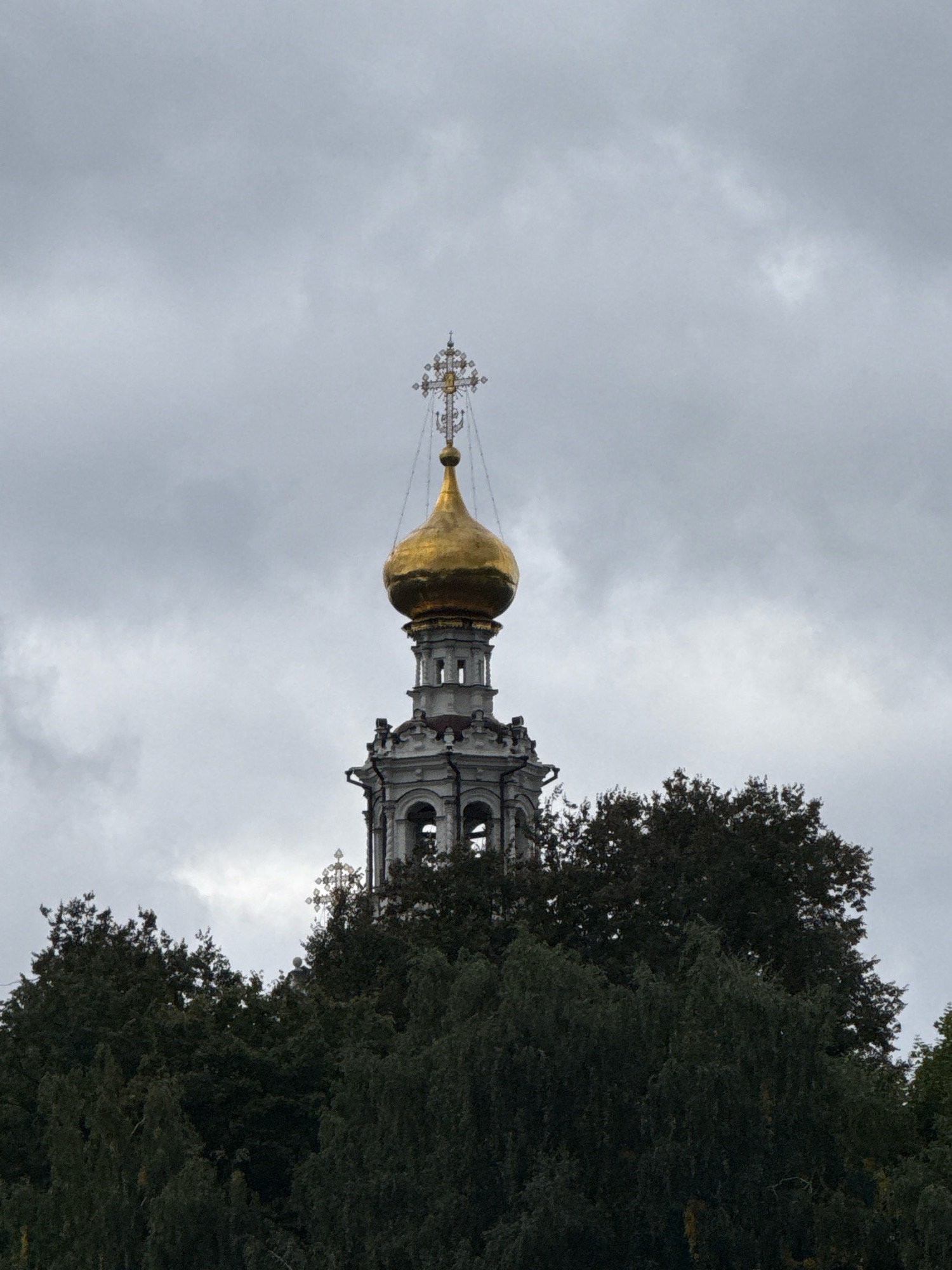 The image shows a close-up shot of the golden dome and cross of a church tower, possibly a bell tower, rising above a dense canopy of dark green trees. The sky is overcast with gray clouds. The architectural style of the tower appears to be ornate and traditional, with white detailing around the belfry. The cross atop the dome is intricate and gilded. The lighting is diffused due to the cloudy weather, casting a muted and somewhat somber tone over the scene. The location is likely a park or a wooded area within Moscow, Russia, given the prevalent presence of church architecture with golden domes in the city.