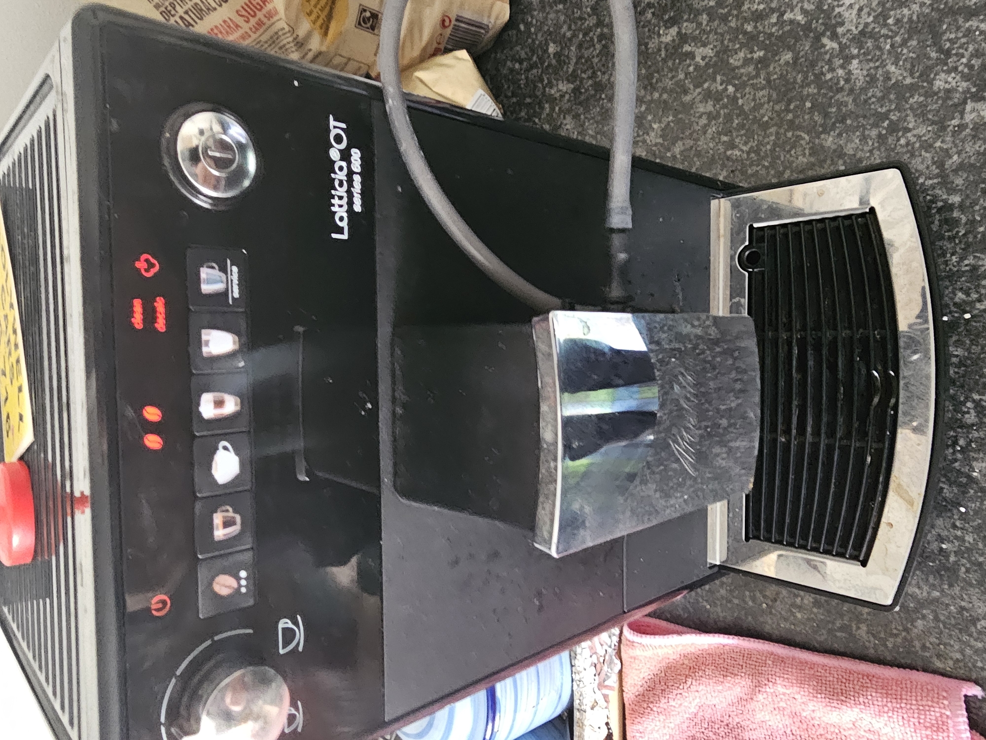 The image shows a Lattissima OT series 600 coffee machine. It is positioned on a gray surface, possibly a countertop. A red and a pink cloth are visible near the base of the machine. In the background, there is a bag of sugar. The coffee machine's control panel is visible, featuring illuminated icons for different coffee types and maintenance functions like cleaning and descaling. The water spout area and the milk frother are also prominent.