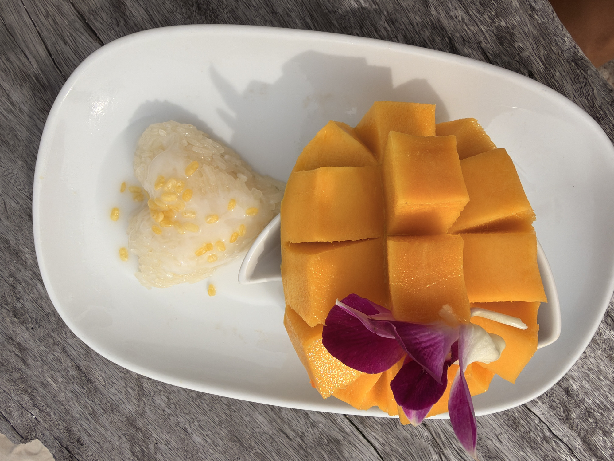 An overhead shot of mango sticky rice on a white oval plate, placed on a weathered wooden surface. The dish features a heart-shaped mound of white sticky rice, topped with a drizzle of creamy white sauce and sprinkled with yellow kernels. Adjacent to the rice is a generous portion of cubed, ripe mango, showcasing its vibrant orange flesh. A delicate purple and white orchid adorns the mango. The wooden background suggests an outdoor setting, possibly a restaurant or cafe in Ko Samui District, Thailand, given the tropical fruit and common dessert presentation.