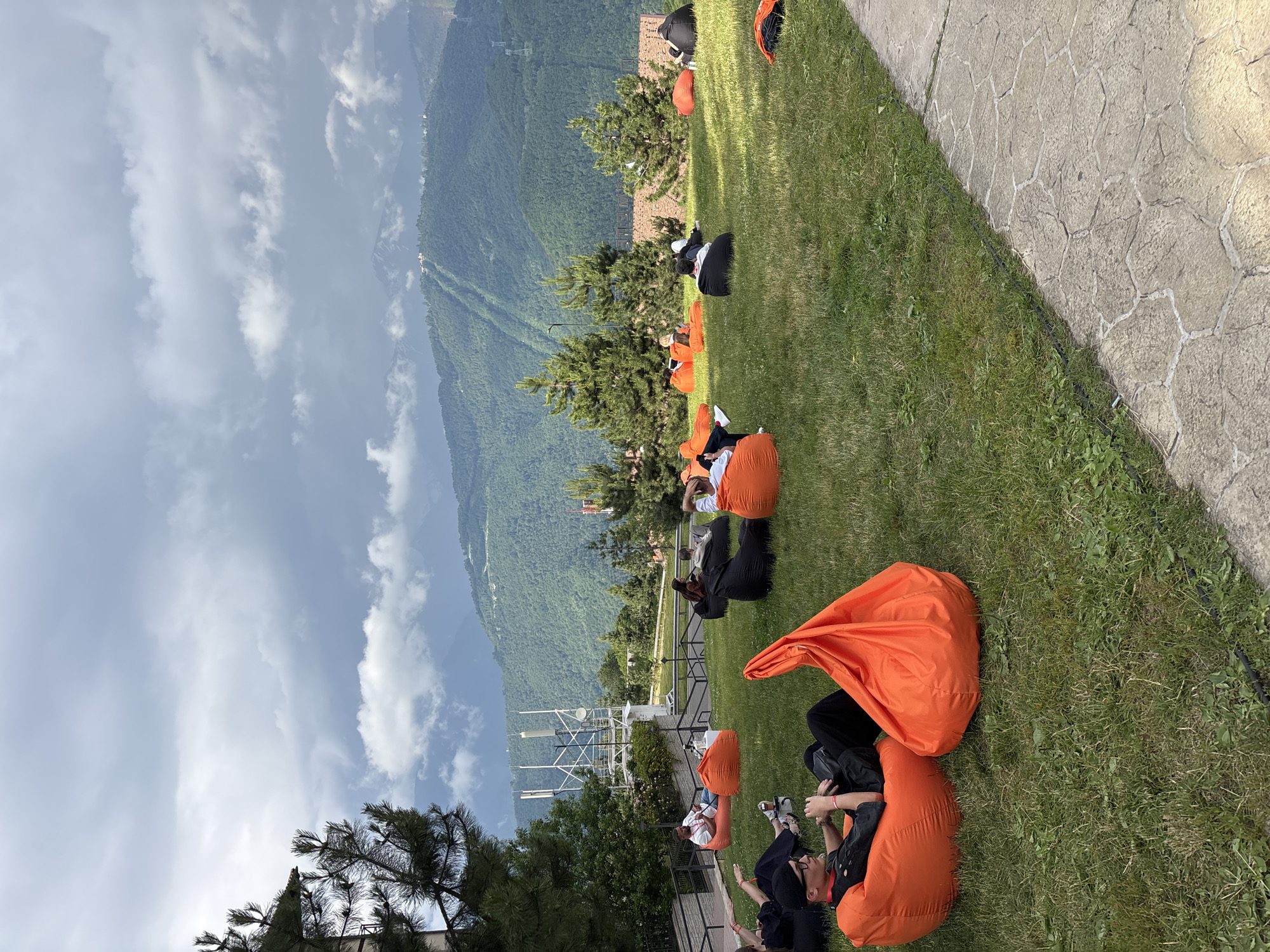 In Estosadok, Russia, a group of people are relaxing on a grassy hillside in front of a scenic mountain range. The people are seated on various beanbag chairs, some orange and some black. One person in the foreground is wearing a black t-shirt, black pants, and a black cap, with glasses perched on their nose. The background features lush green mountains with some cloudy skies and what appear to be ski slopes. A white telecommunications tower with antennas is visible on the left side of the frame, and several evergreen trees are scattered across the hillside. A paved walkway runs along the bottom right of the image.