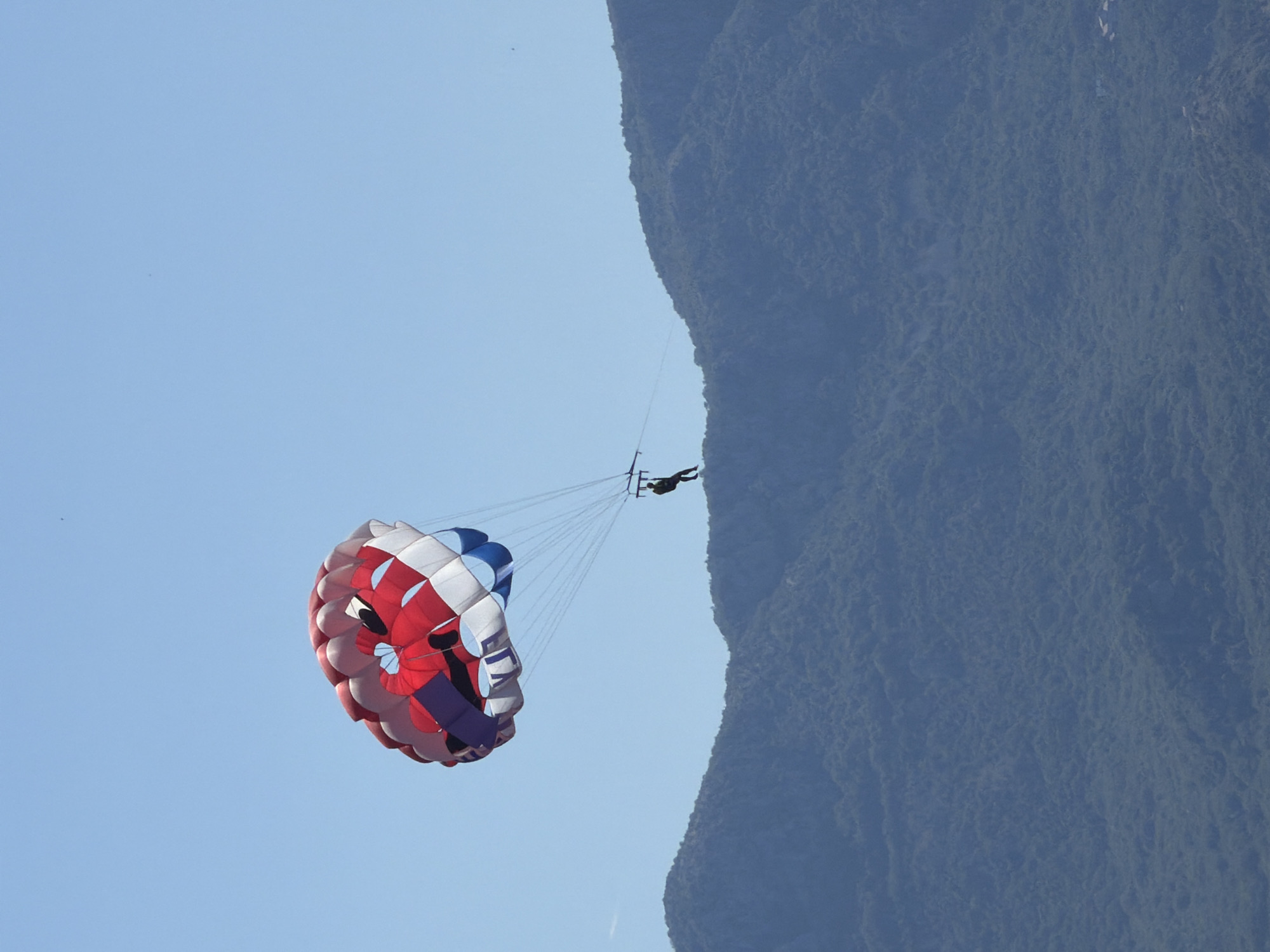 The image shows a person parasailing against a clear blue sky and a backdrop of a steep, forested mountain. The parasailor is suspended in the air, attached to a colorful parasail that features red, white, and blue sections, with a pattern resembling a face. The parasailor appears to be wearing dark clothing and is in a horizontal position, as if gliding.
The scene is set with the bright sun suggesting a daytime activity, likely during fair weather conditions, given the clear sky. The mountain dominates the right side of the frame, its rugged texture and dense vegetation indicating a natural, possibly coastal or lake environment. The presence of the parasailor suggests a recreational or tourist activity taking place.
The overall impression is one of freedom and exhilaration, captured against a dramatic natural landscape. The image is oriented vertically, with the parasail in the foreground and the mountain and sky filling the rest of the frame. There is no visible text in the image.