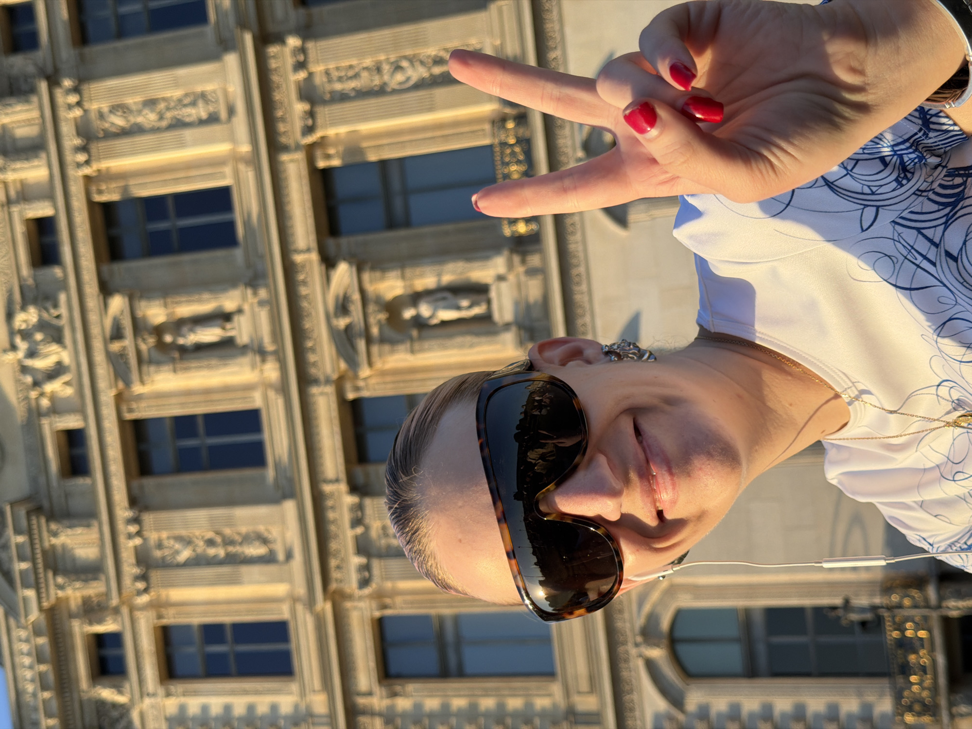 This image, rotated 90 degrees clockwise, presents a close-up selfie of a smiling person against an ornate building in Paris, France. The subject, positioned upside down relative to the background structure, wears large, dark tortoiseshell-patterned sunglasses and a white top adorned with blue swirling patterns. A thin gold chain is visible around their neck, and white wired earbuds are plugged in. The person's left hand is raised, making a "peace" or "V for victory" sign, with fingernails painted in bright red nail polish. A broad, joyful smile is visible.
The background features a grand, light-colored building facade, characteristic of historical Parisian architecture. It is illuminated by warm, golden sunlight, suggesting late afternoon or early morning. The building boasts numerous rectangular windows, deeply recessed, and an abundance of intricate decorative elements. These include detailed carvings, possibly small statues or bas-reliefs, and elaborate moldings that frame the windows and columns. The strong highlights and shadows indicate a clear, sunny day. The overall impression is a cheerful moment captured against a beautiful, historic urban backdrop. No text is visible in the image.