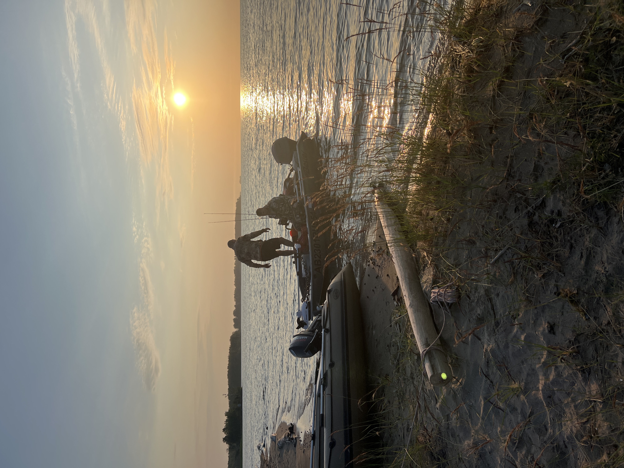 The image depicts a serene sunset scene on a sandy shore, likely located in Borovoe, Russia, given the context. Two individuals, silhouetted against the vibrant orange sky, are standing in or near an inflatable boat. One person is visible from the back, stepping into the boat, while the other appears to be already seated. Another inflatable boat is resting on the shore to the left, partially submerged. The water is calm, reflecting the warm hues of the setting sun, with a gentle ripple effect on the surface. Tall grasses and a piece of driftwood line the foreground, adding to the natural, rustic ambiance. The overall atmosphere is peaceful and tranquil, capturing a moment of outdoor activity at dusk.