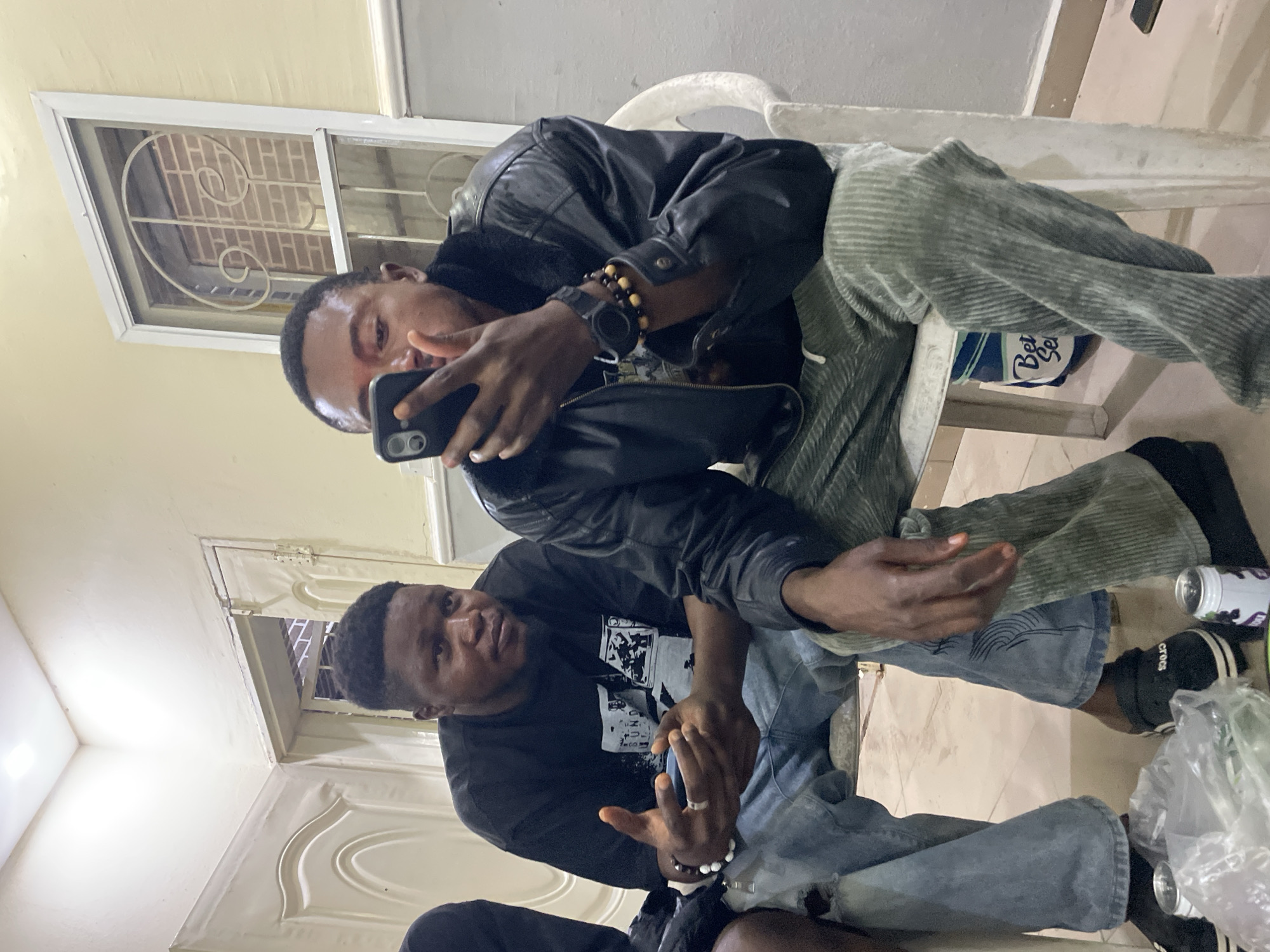 Two African men are sitting in chairs indoors. The man at the top is wearing a black leather jacket, a watch, and a bracelet. He is holding a black smartphone and looking at the screen. The man at the bottom is wearing a black T-shirt and jeans, also with a bracelet. The environment appears to be a simple room with tiled floors and white walls. There is a window in the background, and general lighting suggests an interior setting. A bottle and some plastic bags are visible near their feet. The location is likely Jos, Nigeria, based on the prompt.