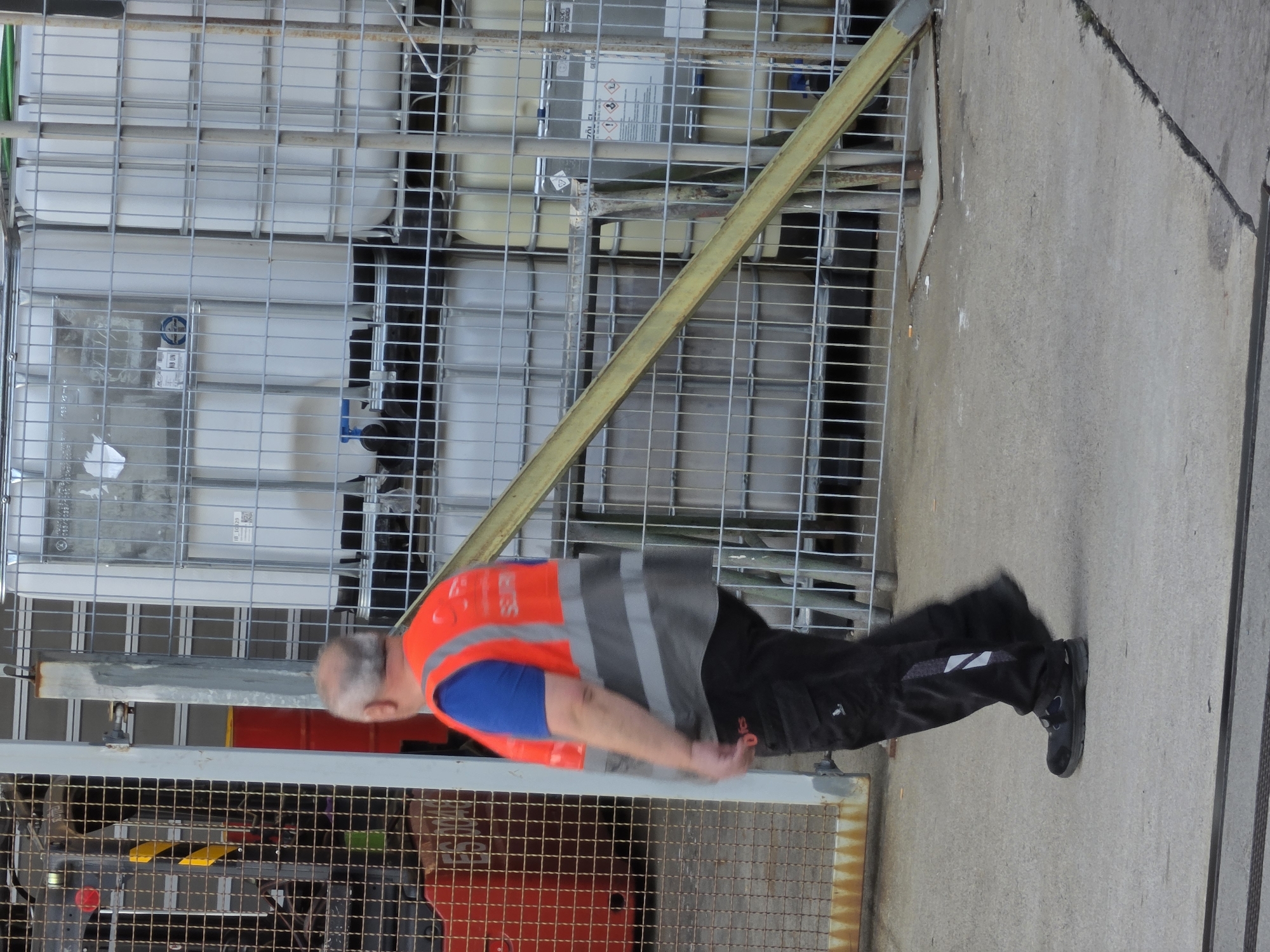 A man wearing an orange and gray high-visibility vest over a blue t-shirt and black pants walks on a concrete surface in Walldorf, Germany. He is wearing dark shoes. Behind him, a chain-link fence encloses large white plastic containers, some with blue valves. To the left, a red barrel is visible behind another section of fence. The background shows a metallic structure.