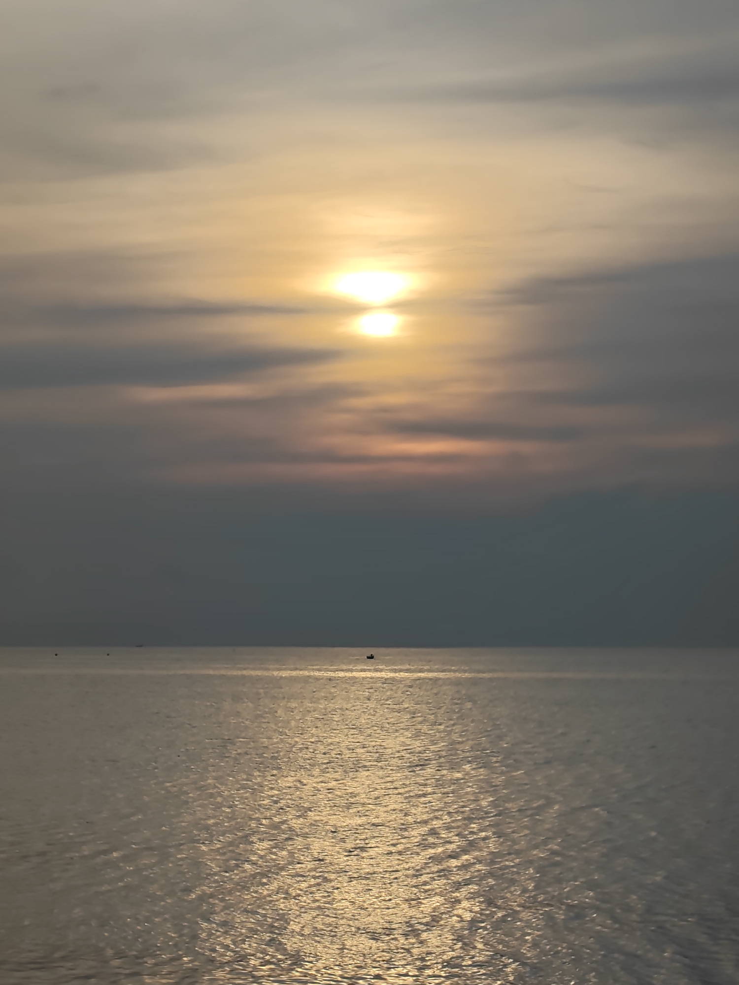 The image depicts a sea at sunset, possibly in Lhokseumawe, Indonesia, given the prompt. The sun is positioned in the center of the frame, partially obscured by clouds. The water surface reflects the sunlight, creating a bright path towards the viewer. A few small boats are visible on the horizon. The sky is a mix of grey and golden hues. The image conveys a sense of calm and vastness.