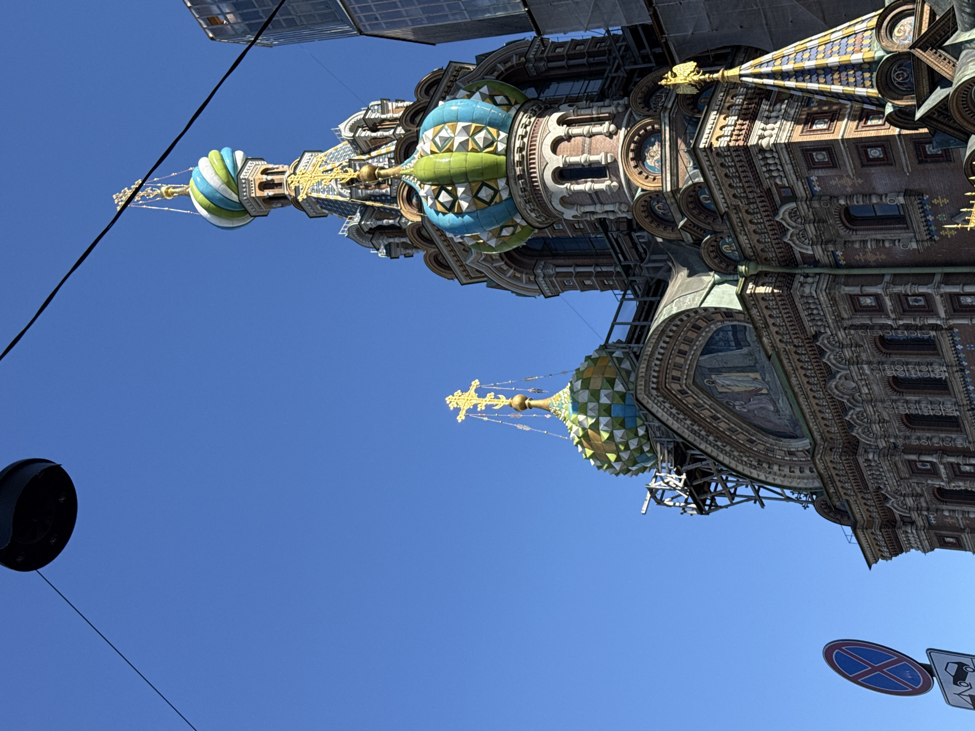 This media file is a low-angle shot of the exterior of a highly ornate, multi-domed building, likely a church, under a clear, bright blue sky. The building's architecture is characterized by intricate detailing, colorful tiled domes, and golden embellishments, including crosses atop the spires. One of the domes is predominantly blue and green, with a patterned surface, and a golden cross at its apex. Another dome, more distant, also features green and blue tiling. The building appears to be undergoing some form of construction or renovation, as scaffolding is visible around some sections.
In the foreground, a black, circular object, possibly a light fixture or camera, hangs from a thin black wire that stretches across the frame. Another wire is visible in the upper left portion of the image.
In the lower right corner, two traffic signs are partially visible. One is a blue oval sign with a red cross or 'X' inside. The other is a white rectangular sign with black symbols, one of which resembles a tow-away symbol. The presence of these signs suggests an urban setting.
The bright sunlight and clear sky indicate daytime with fair weather. The orientation of the image is rotated, with the building's architectural elements appearing to extend upwards. The overall impression is one of grandeur and historical significance, juxtaposed with modern elements like the wires and traffic signs. No people are visible in the image. The location is identified as Saint Petersburg, Russia, which aligns with the architectural style commonly found in the city.