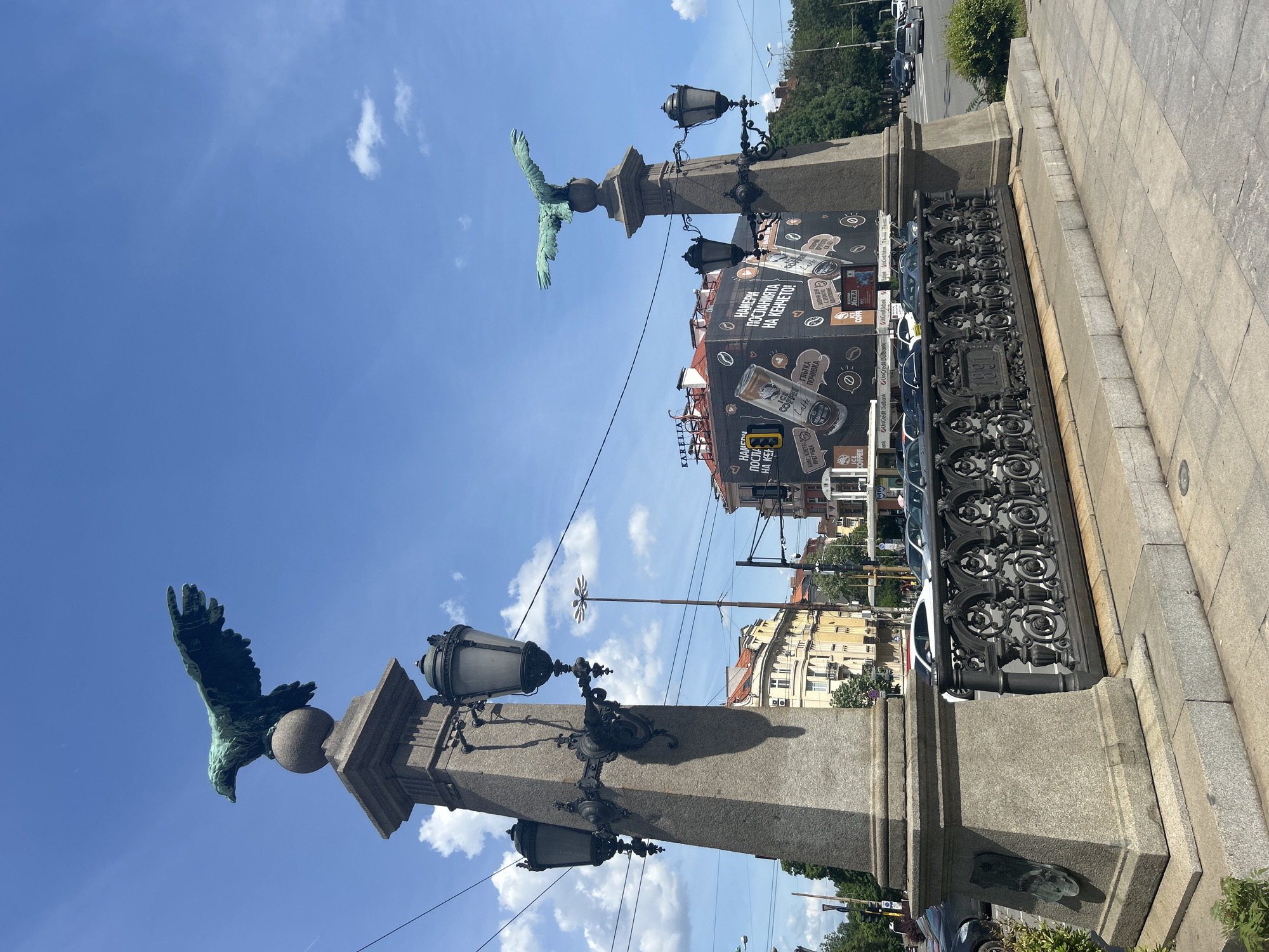 The image shows a street scene in an urban area, likely a bridge or elevated walkway, marked by ornate stone pillars topped with bronze sculptures of eagles. Each pillar is also adorned with decorative lamp fixtures. Between the pillars, there is a wrought iron railing. A large advertisement is affixed to the railing or a structure behind it, featuring images of coffee products and Bulgarian text, indicating the city might be located in Bulgaria. Cars are parked along the street, and buildings of varying heights and architectural styles can be seen in the background. A utility pole stands next to a white and silver wind turbine like decoration on a pole. The sky is blue with scattered clouds, suggesting it is daytime and the weather is clear. The pavement is made of rectangular stone slabs.