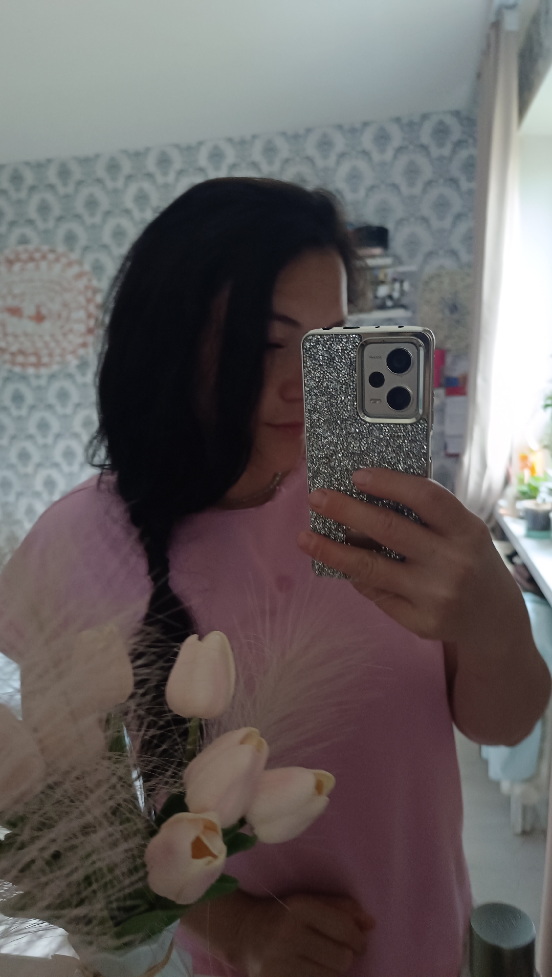 The image is a mirror selfie taken indoors, likely in a residential setting. A person with long, dark hair is holding a smartphone with a sparkly silver case, pointed towards the mirror. The person is wearing a pale pink t-shirt. In the foreground, a bouquet of white and pale pink tulips is visible. The background shows a wall with patterned wallpaper and a window with sheer curtains. There are no explicit indicators of the city of Chaikovsky, Russia, in the image.