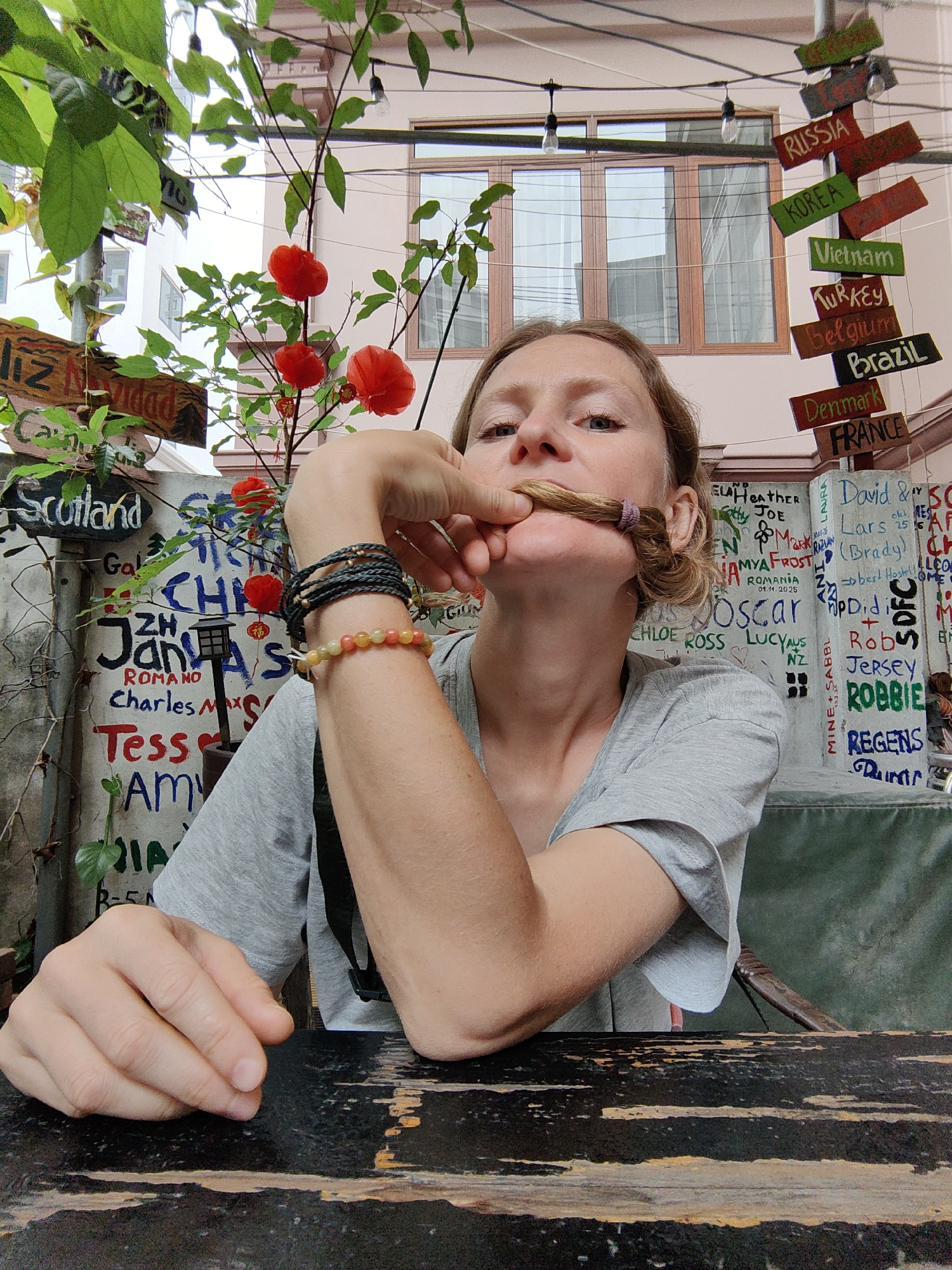 The image is a selfie taken in what appears to be an outdoor seating area of a cafe or restaurant. The location is in Bai Chay Ward, Vietnam, evidenced by a signpost indicating different countries, including Vietnam. A fair-skinned woman is the primary subject. She is wearing a gray T-shirt and has her hair pulled up, with a section of it held in front of her mouth as if creating a makeshift mustache. She has bracelets on her left wrist.
The background includes a wall covered in graffiti-like writings of names and dates, as well as plants with red flowers. Additional signs display place names such as "Scotland" and "Liz Navidad". A dark-colored, aged wooden table is in the foreground. Overhead, strings of lights and more signs (including "Russia", "Korea", "Vietnam", "Turkey", "Belgium", "Brazil", "Denmark", and "France") suggest a layered and decorated outdoor space. A building facade with windows is visible in the background.