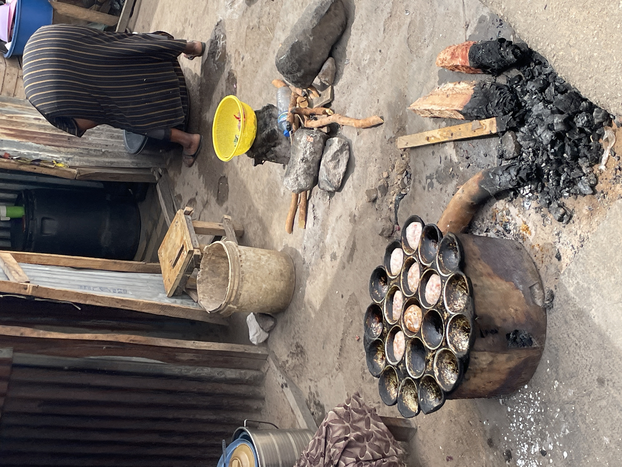 The image shows an outdoor cooking scene in Jos, Nigeria. A person wearing a striped garment bends over next to a corrugated metal wall, near a yellow plastic basket and a bucket. A stove-like contraption with multiple circular depressions filled with light-colored food sits nearby, connected to a pipe leading to a pile of burning wood and charcoal. Stones and wood are arranged around the fire. A wooden stool is visible near the bucket. Other containers and items are scattered on the ground, which appears to be a concrete surface.
