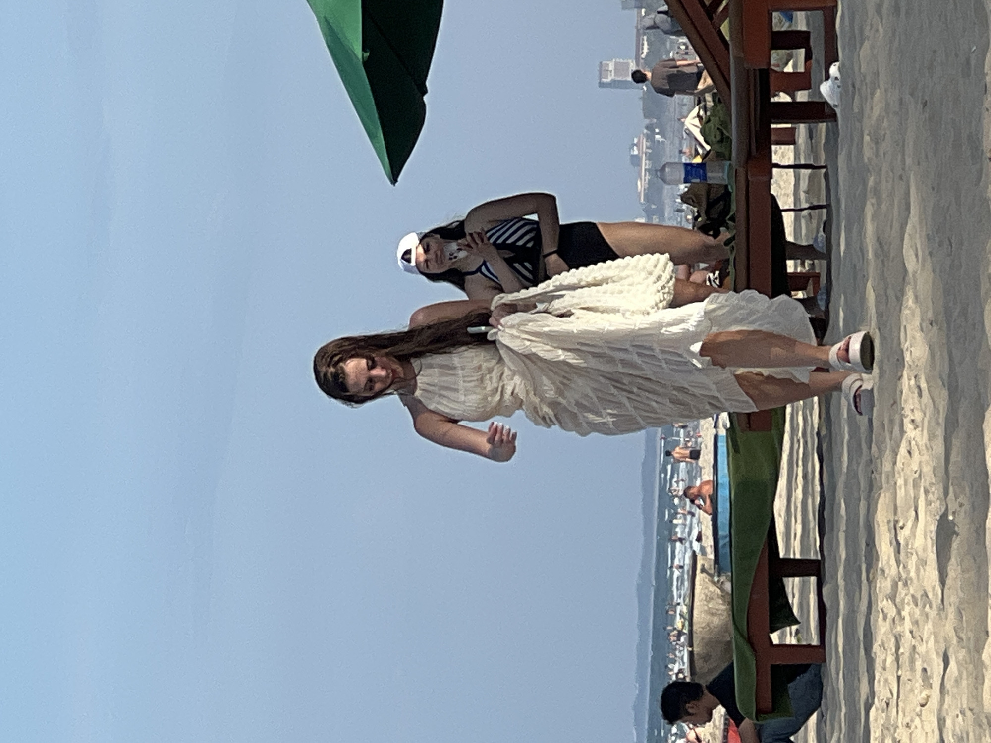 On a sandy beach in Da Nang, Vietnam, a woman in a flowy white dress walks towards the camera. She is holding a white bag and a piece of fabric. Behind her, another woman in a black and white striped swimsuit, wearing a white baseball cap, holds a phone and appears to be taking a photo. The background shows a crowded beach with people in the water and on the sand, a green umbrella, lounge chairs, and buildings in the distance. The sky is clear and blue.